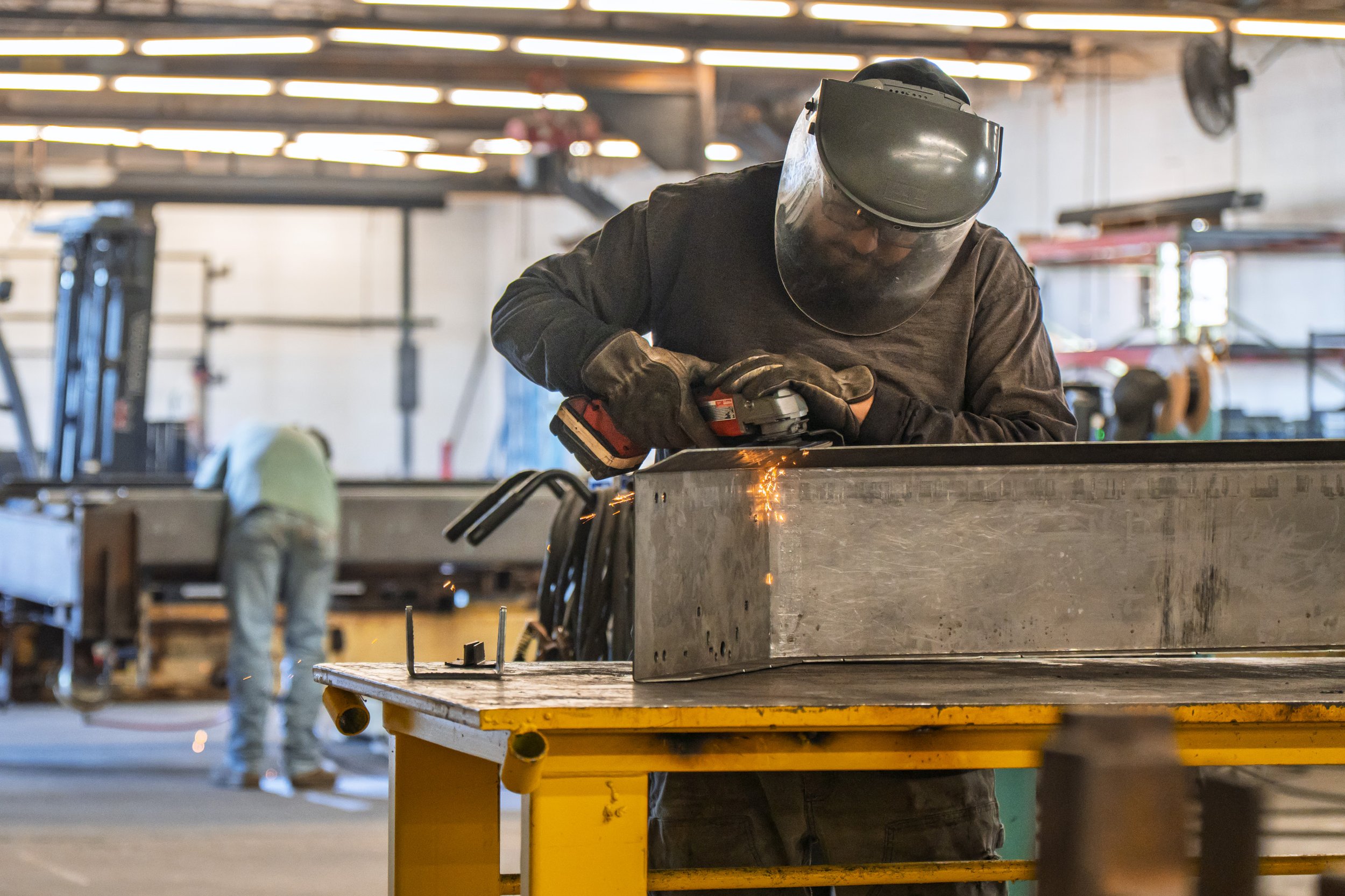 A man wearing a welding helmet and gloves is using a power tool to weld a large metal piece inside a manufacturing workshop. Sparks are visible as he works.