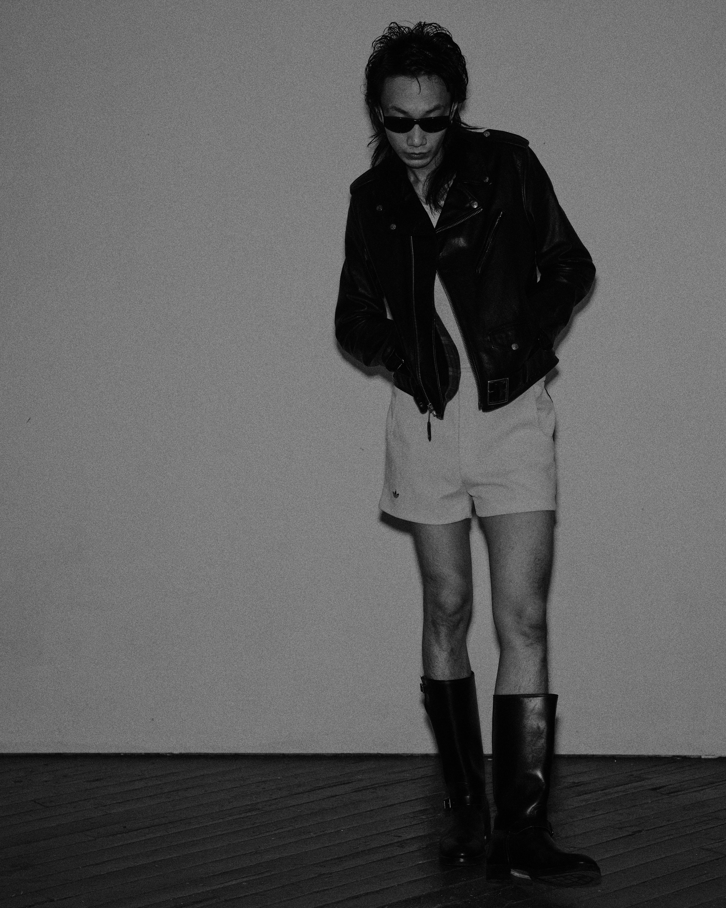 A person wearing a leather jacket, shorts, tall boots, and sunglasses standing against a plain background.