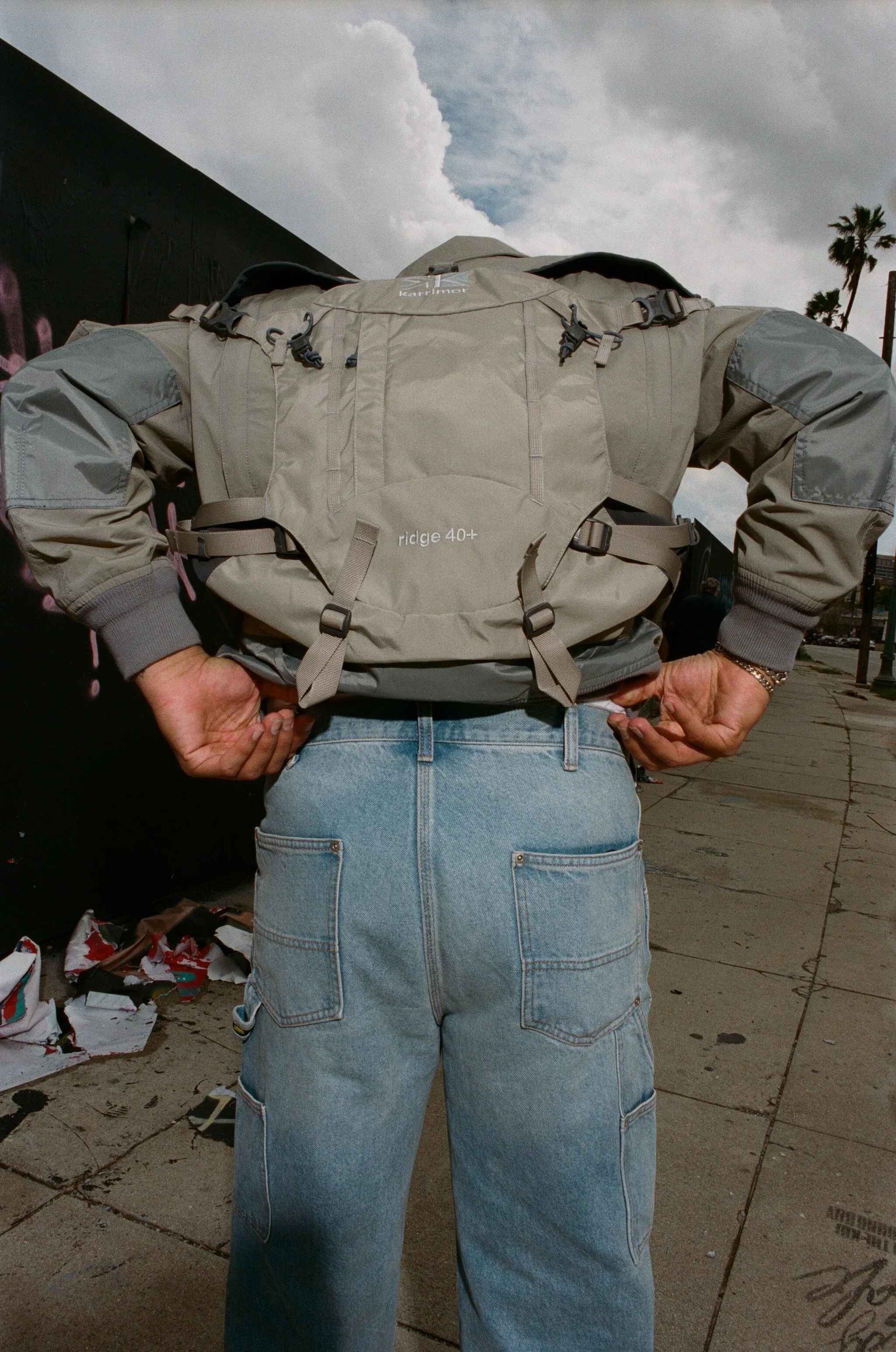 Person standing on sidewalk with hands on hips, wearing a gray backpack and light blue jeans, with a cloudy sky and palm trees in the background.