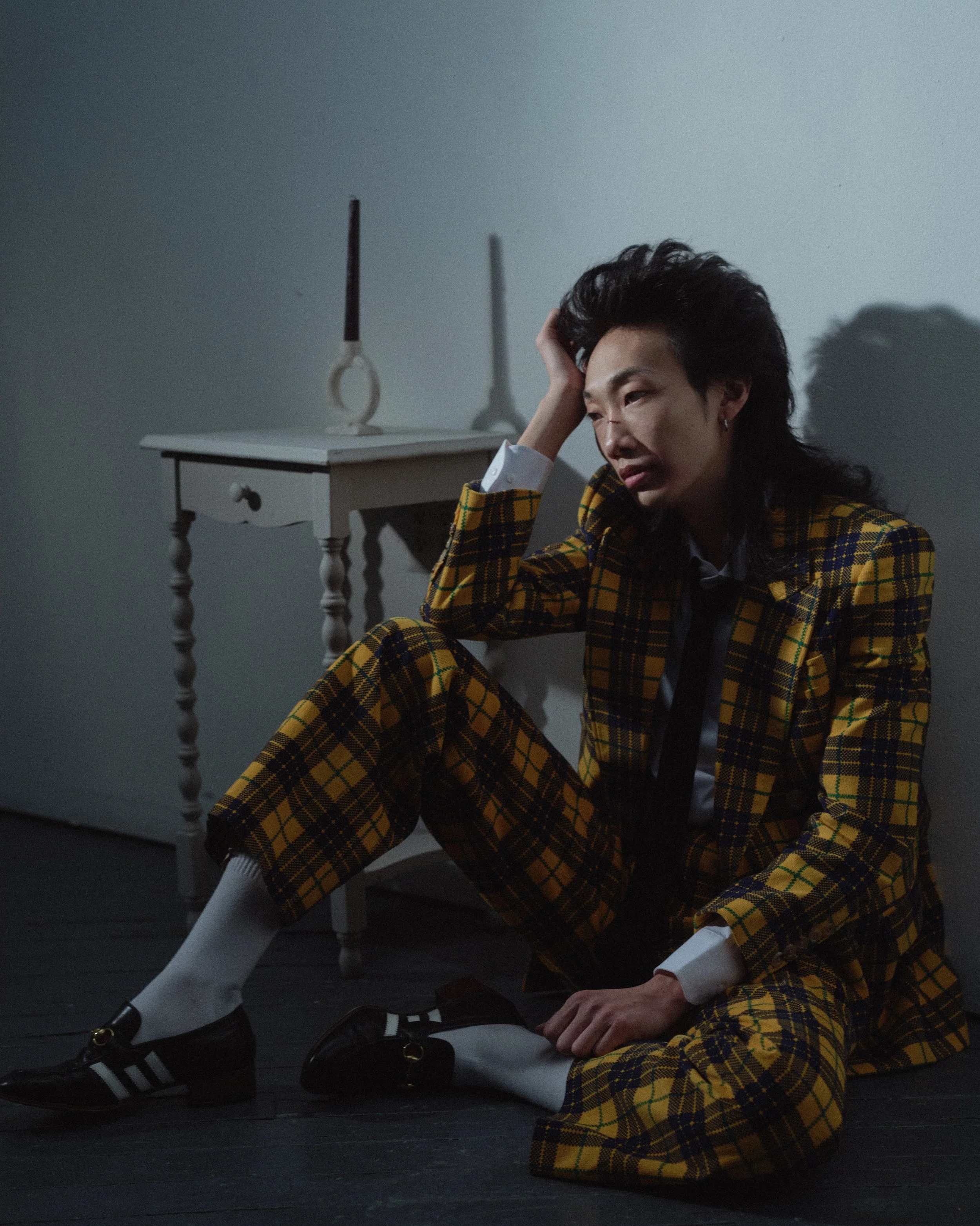 Person sitting on the floor wearing a yellow plaid suit, black shoes, and white socks, with a distressed expression, resting head on hand next to a white table with candles on it.