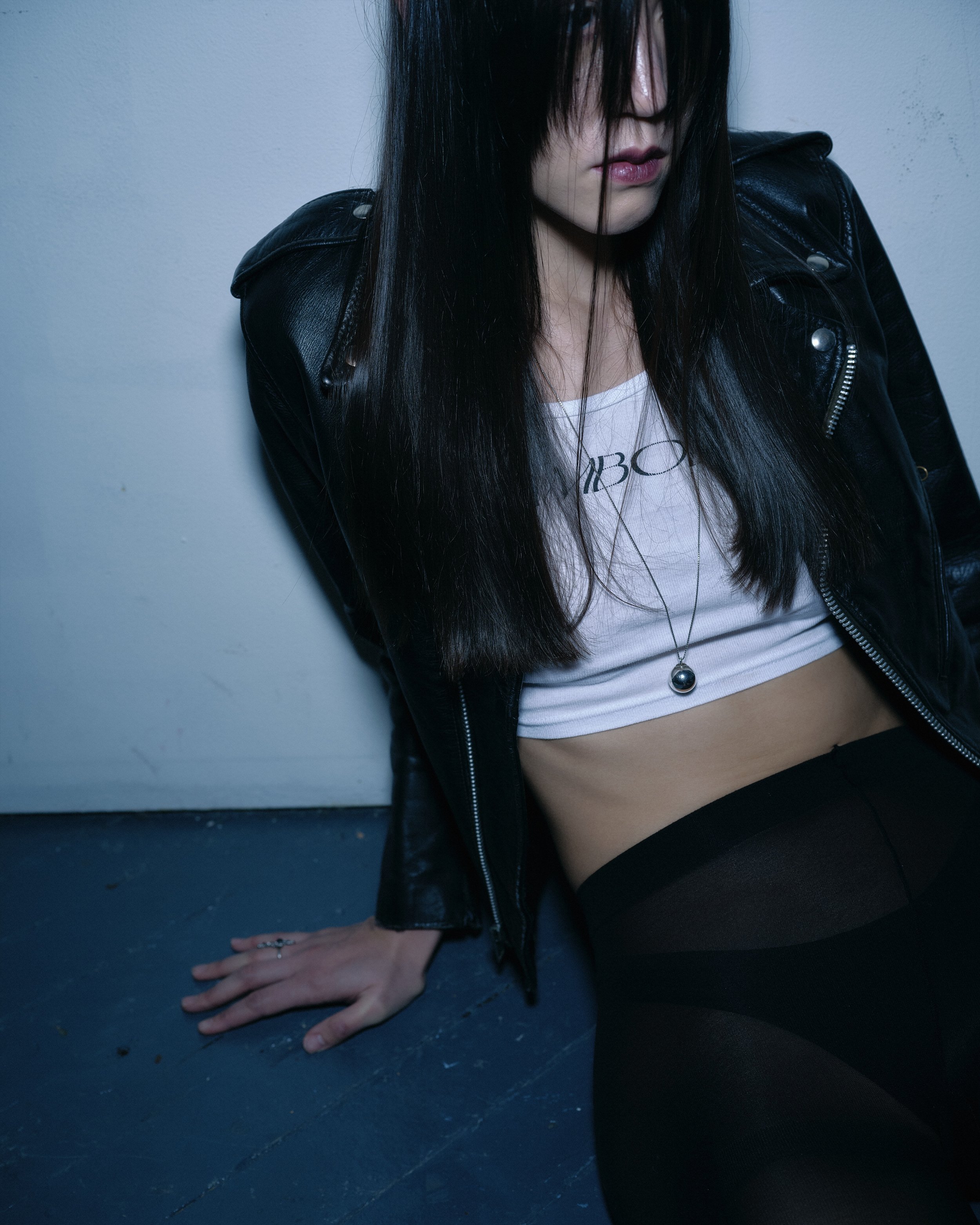 A woman with long black hair and fair skin sitting on the floor against a white wall, wearing a black leather jacket, a white crop top, black leggings, a necklace, and a ring.