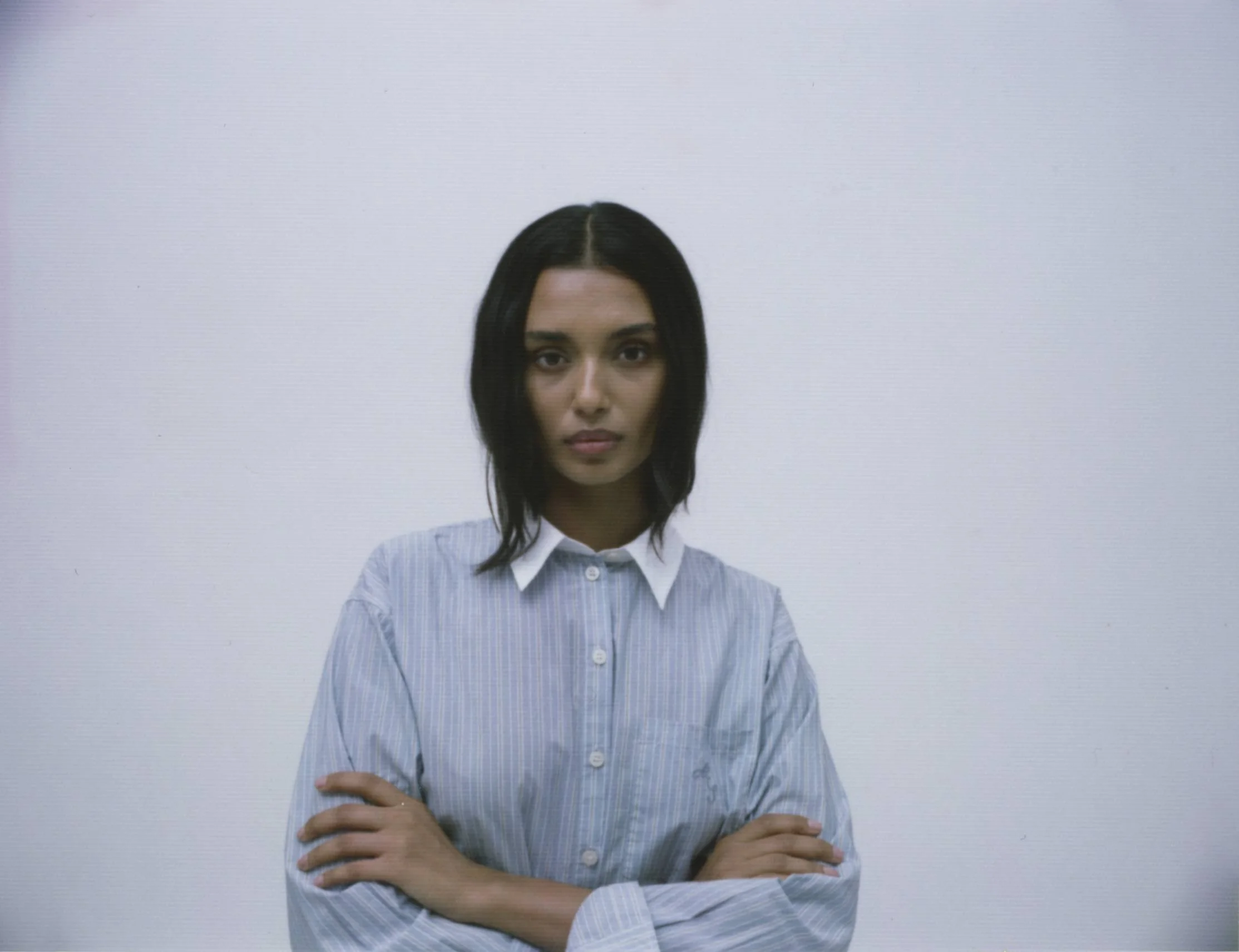 A young woman with dark hair in a shoulder-length style wearing a blue and white striped button-up shirt with arms crossed against a plain white background.