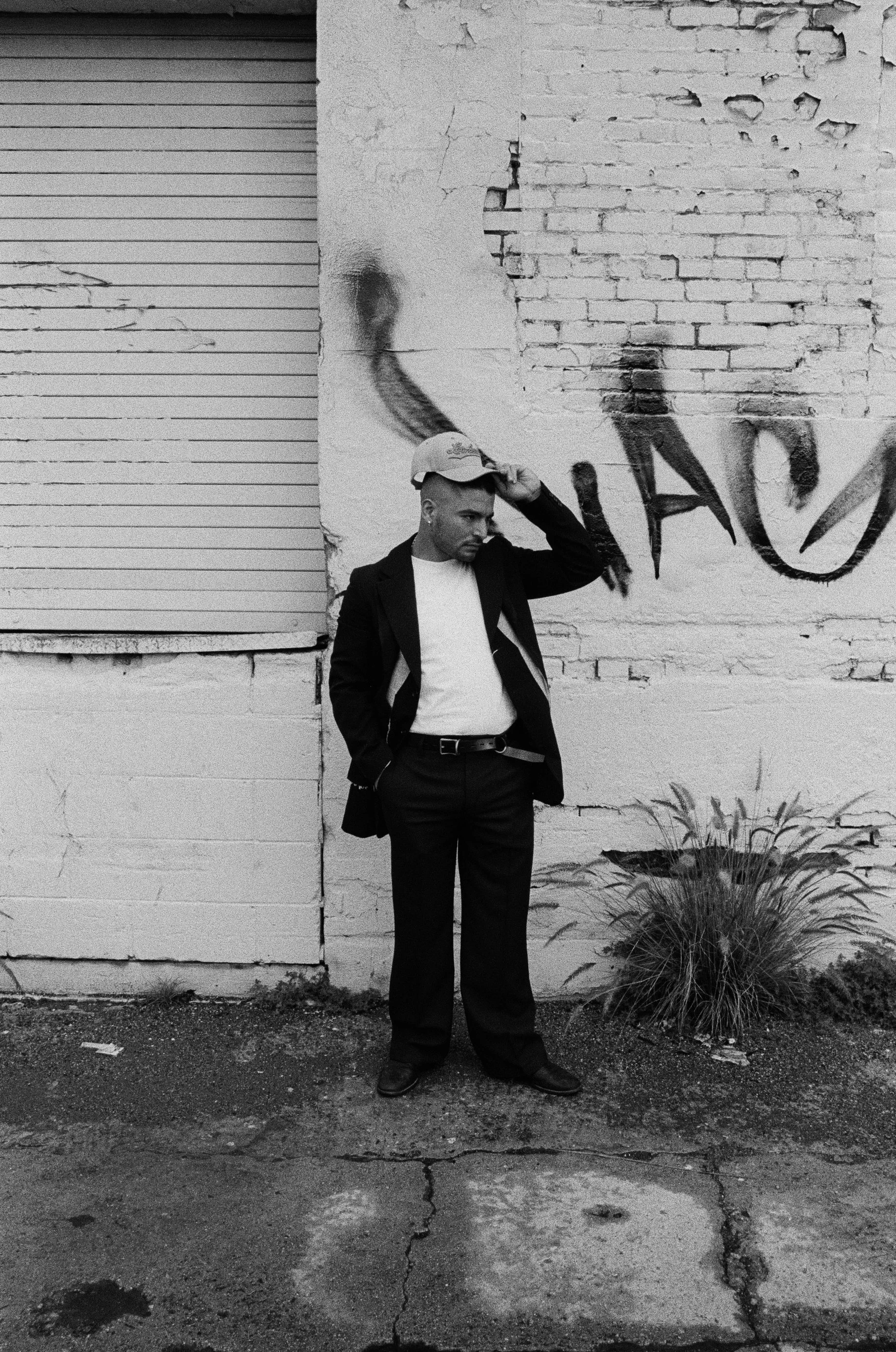 A man in a suit and cap standing against a brick wall with graffiti, looking down with hand on his cap.