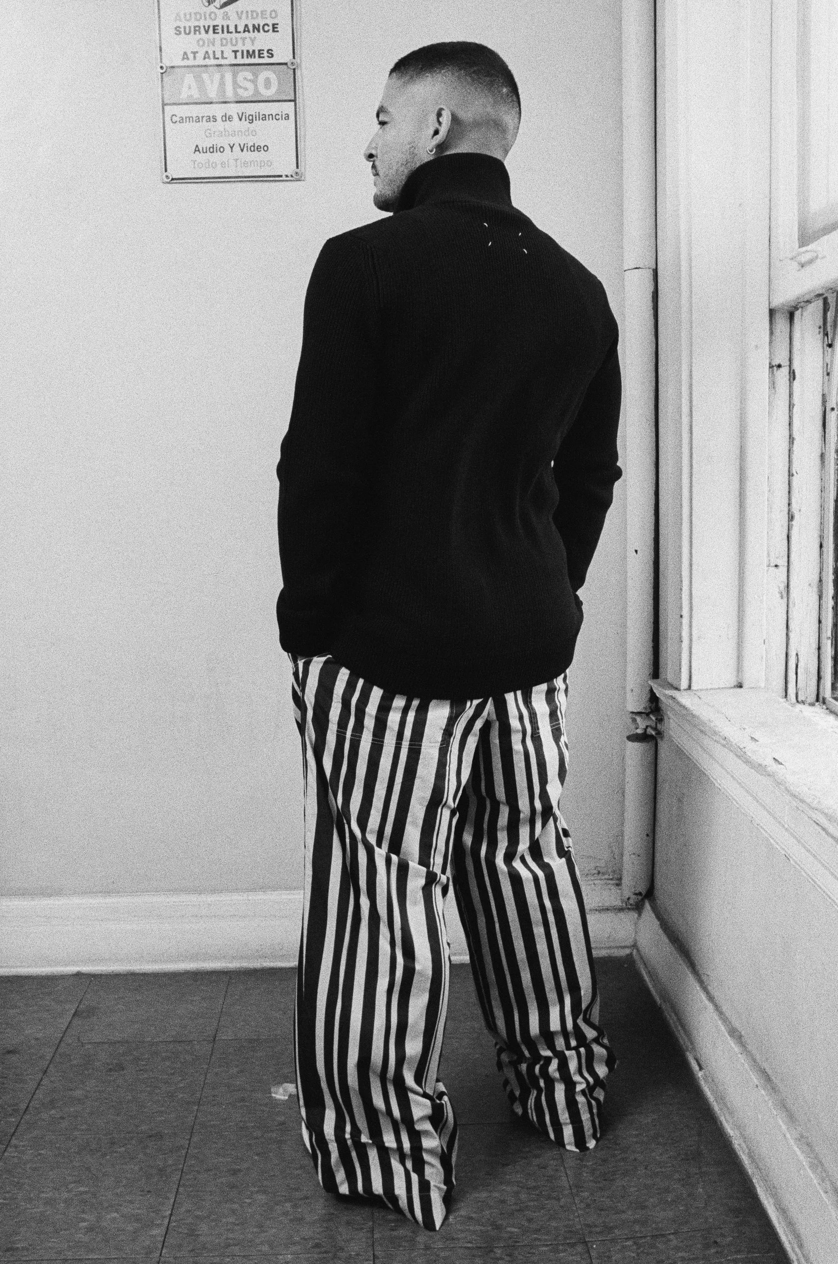 A man with short hair and a beard standing indoors, wearing a black turtleneck sweater and black and white vertically striped pants. He is facing sideways near a corner window with signs on the wall behind him.