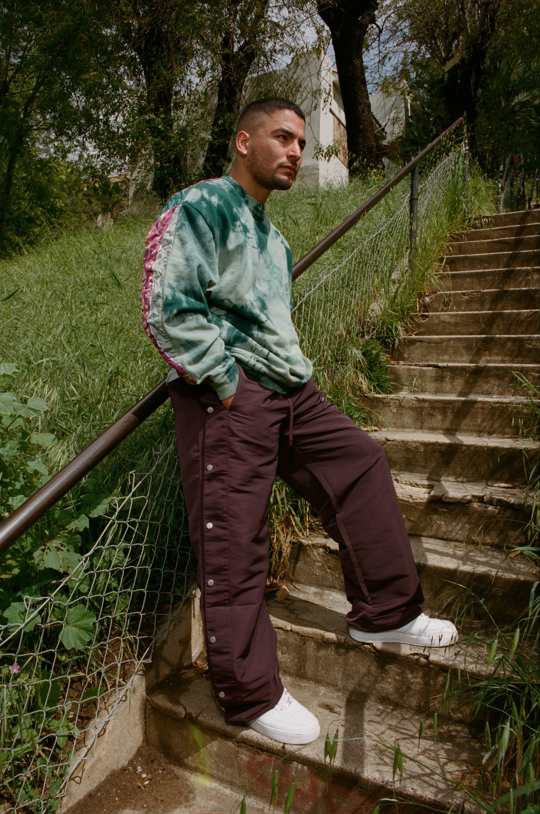 A young man leaning against a railing on outdoor concrete stairs, wearing a tie-dye sweatshirt, burgundy pants, and white sneakers, with greenery and trees in the background.