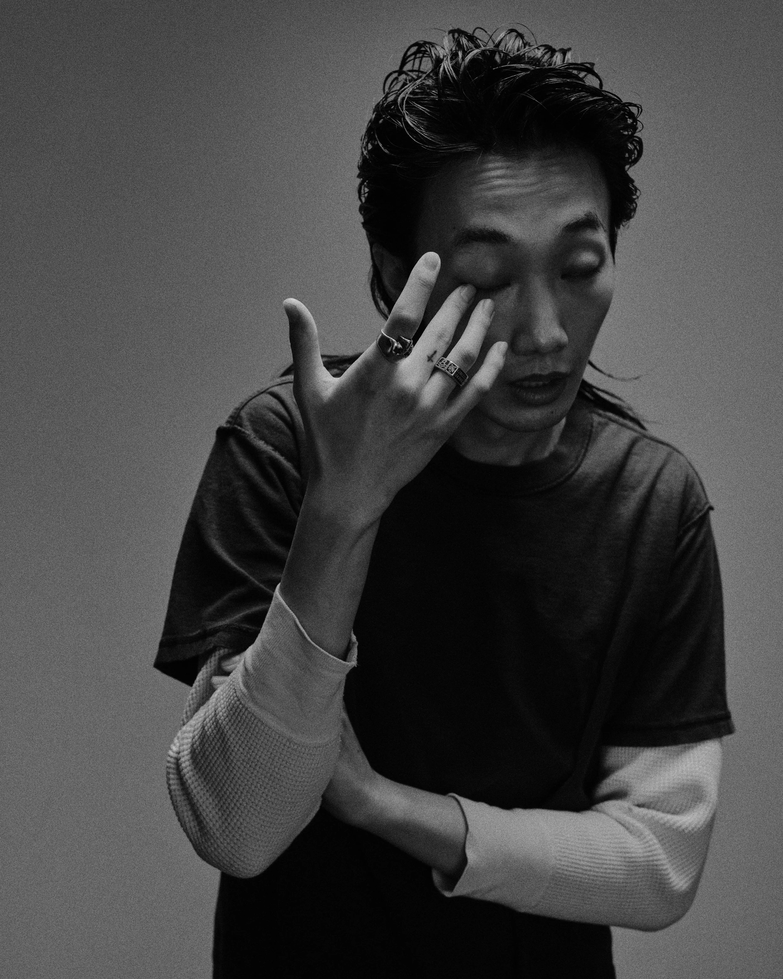 Black and white photo of a person with wavy hair, wearing rings and a layered shirt, touching their eye with one hand, with a neutral background.