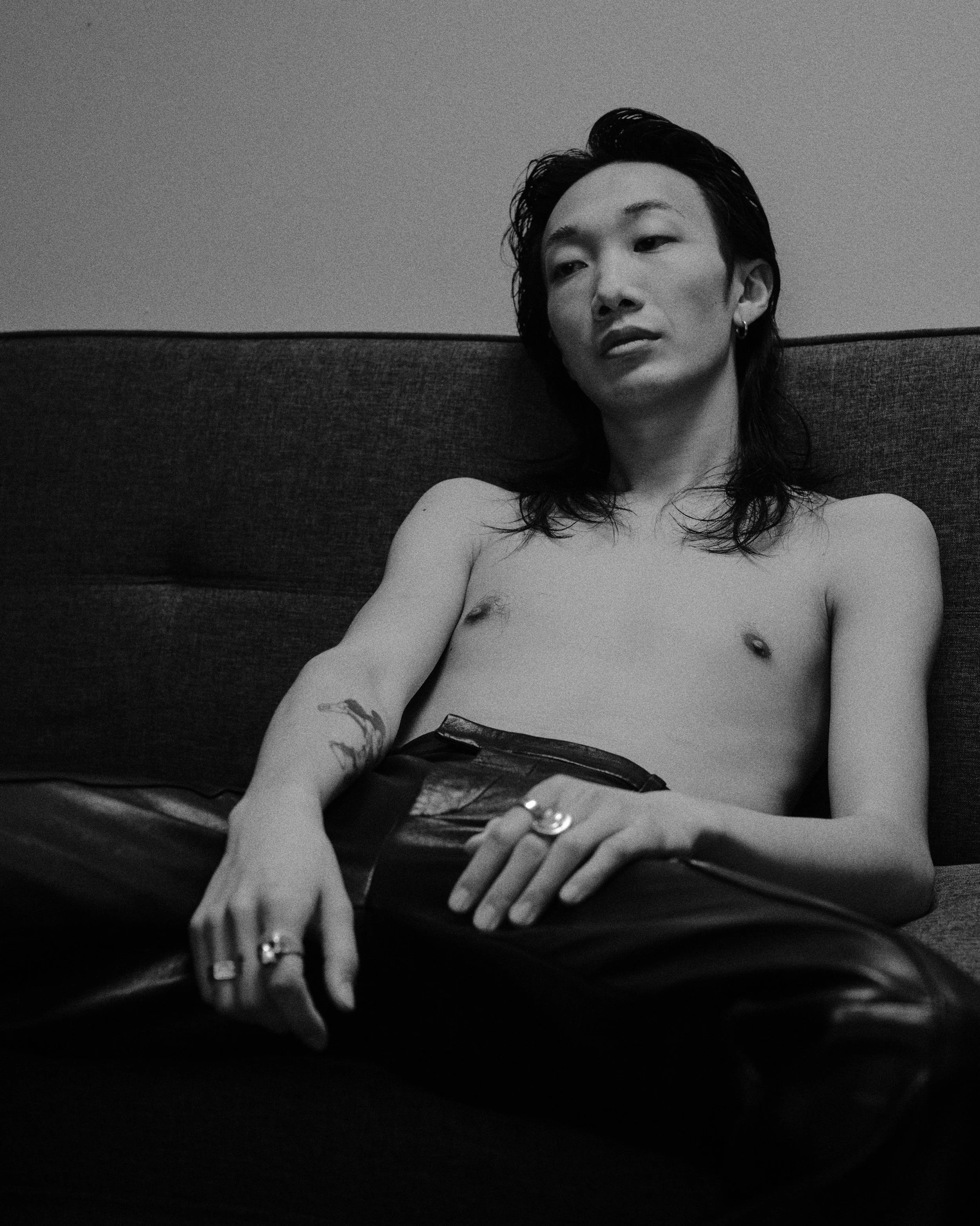 Black and white photo of a shirtless person with long hair, sitting on a dark couch, wearing black leather pants and rings, with tattoos on their arm, looking to the side.