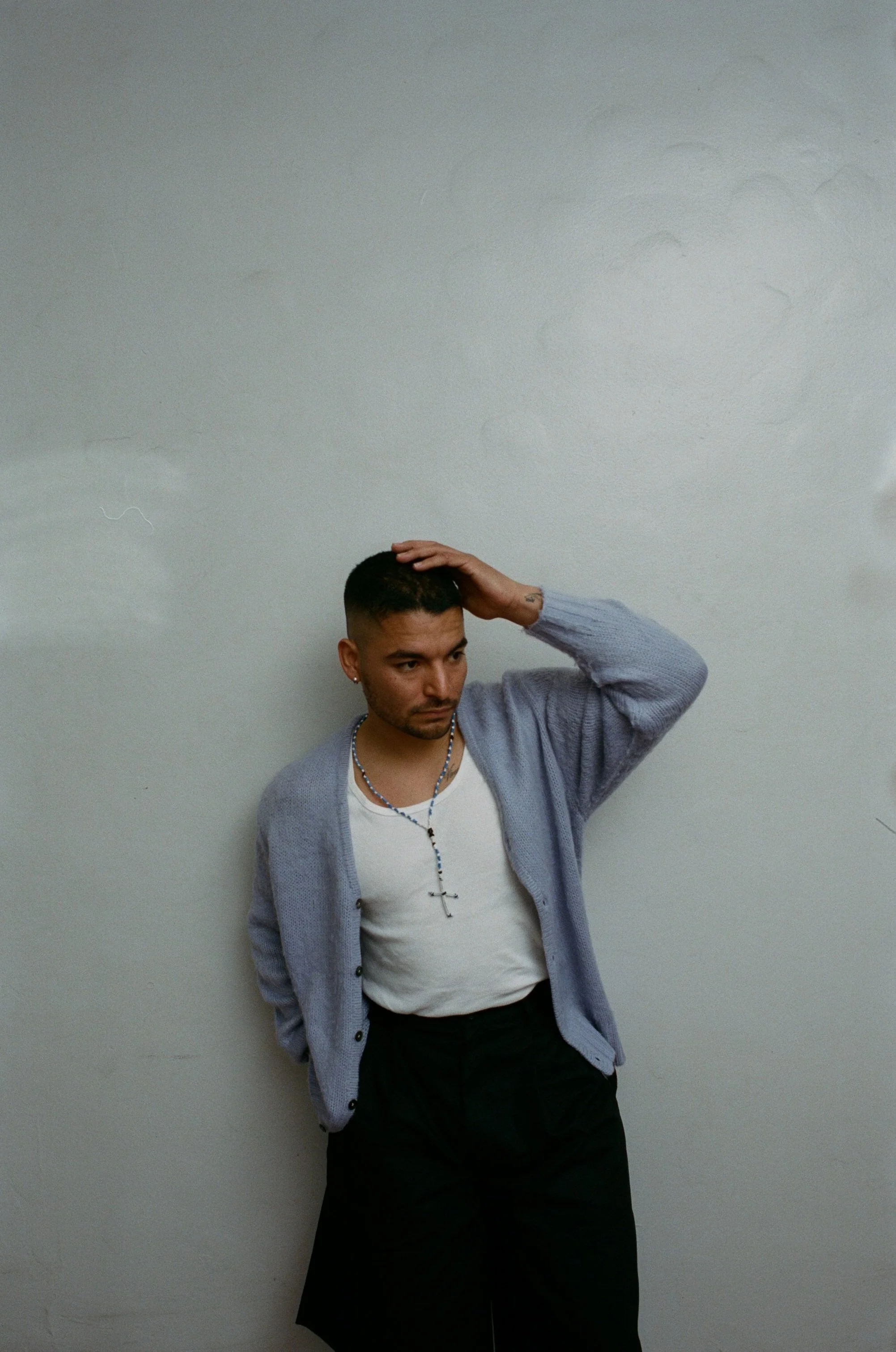 A man with short dark hair and hoop earrings, wearing a white t-shirt, a grey cardigan, and a rosary necklace, stands against a plain light-colored wall with his left hand resting on his head and looking down.