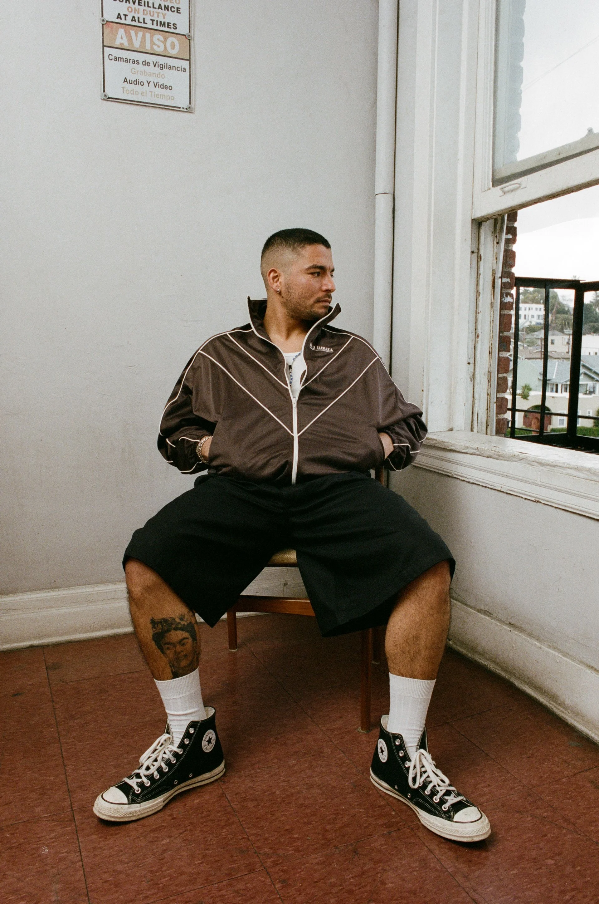 A man with short hair and a beard sitting on a chair near a large window, wearing a brown jacket, black shorts, white socks, and black Converse sneakers, with a tattoo on his left calf of a woman's face.