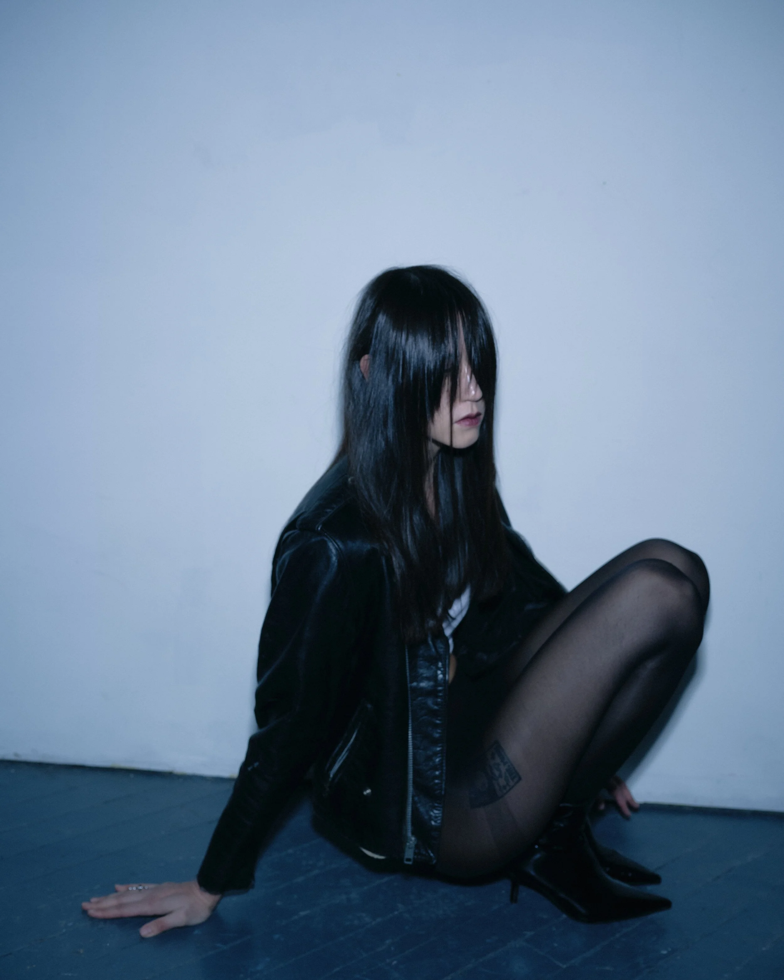 A woman with long, dark hair sitting on the floor against a plain wall, wearing a black leather jacket, thigh-high black stockings, and black boots.