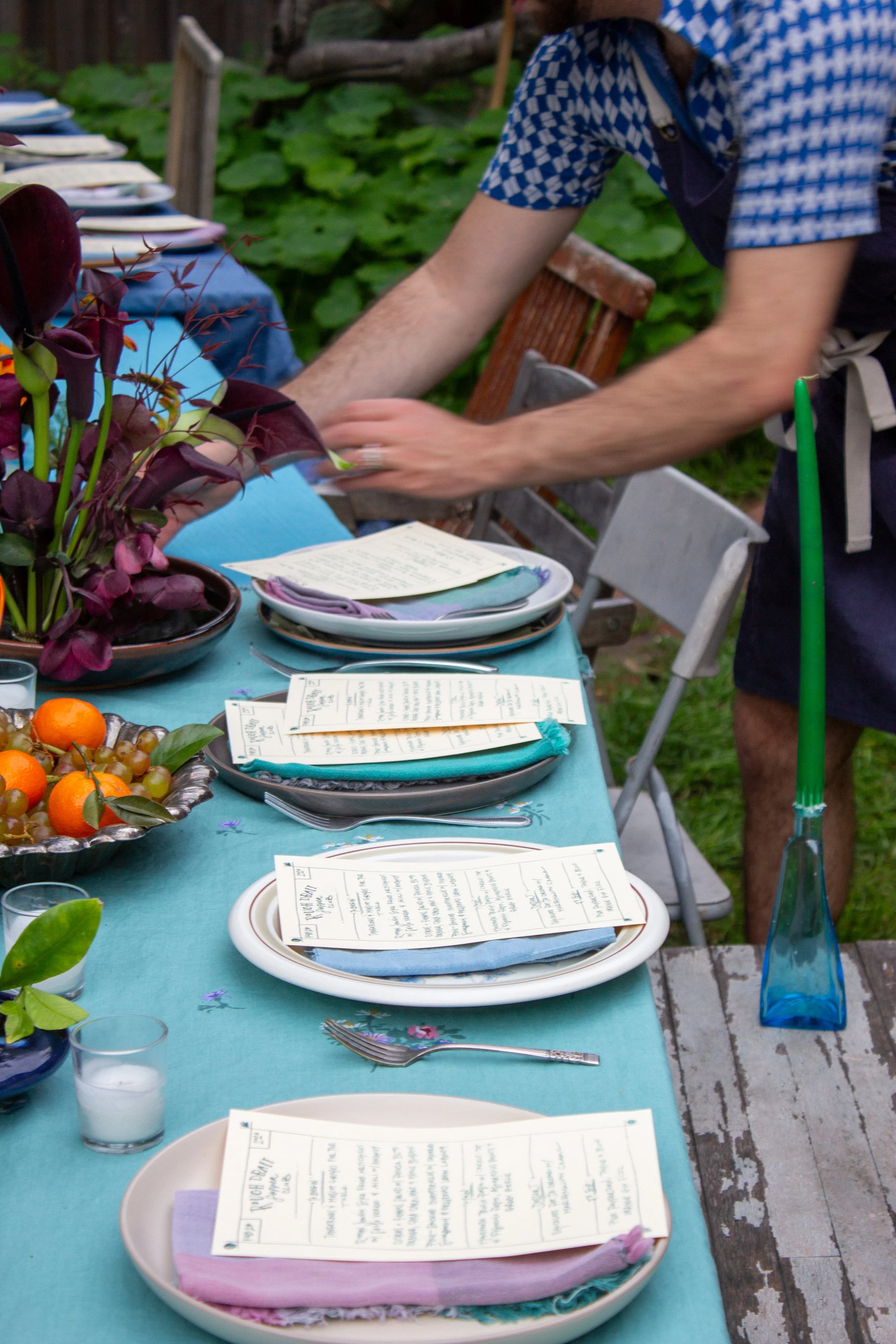 A table set for a meal outdoors, with plates, napkins, menus, glasses, candles, fruit, and decorative flowers.