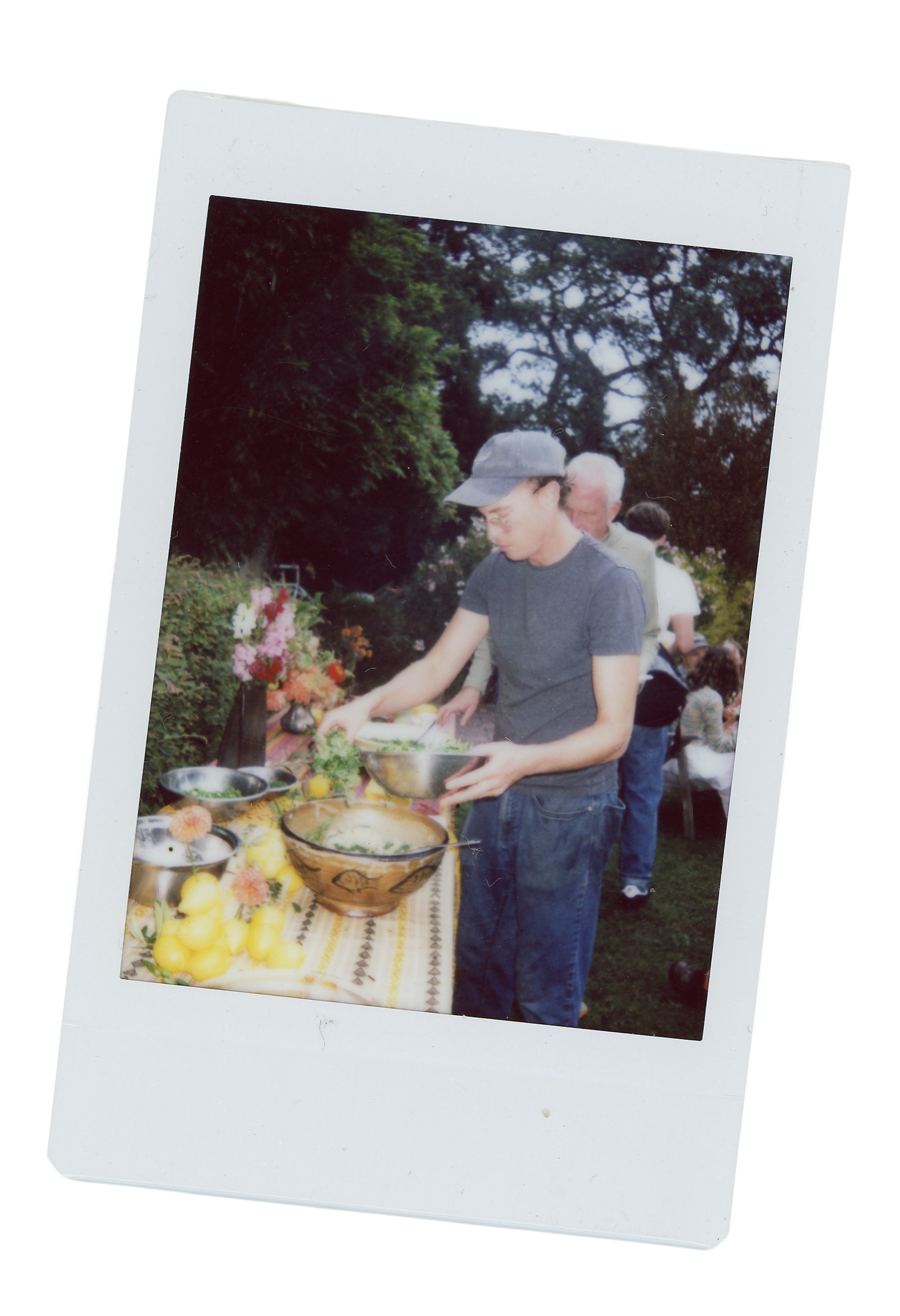 Young man in a gray cap and T-shirt preparing food at an outdoor table with fruits and flowers, surrounded by people in a garden setting.