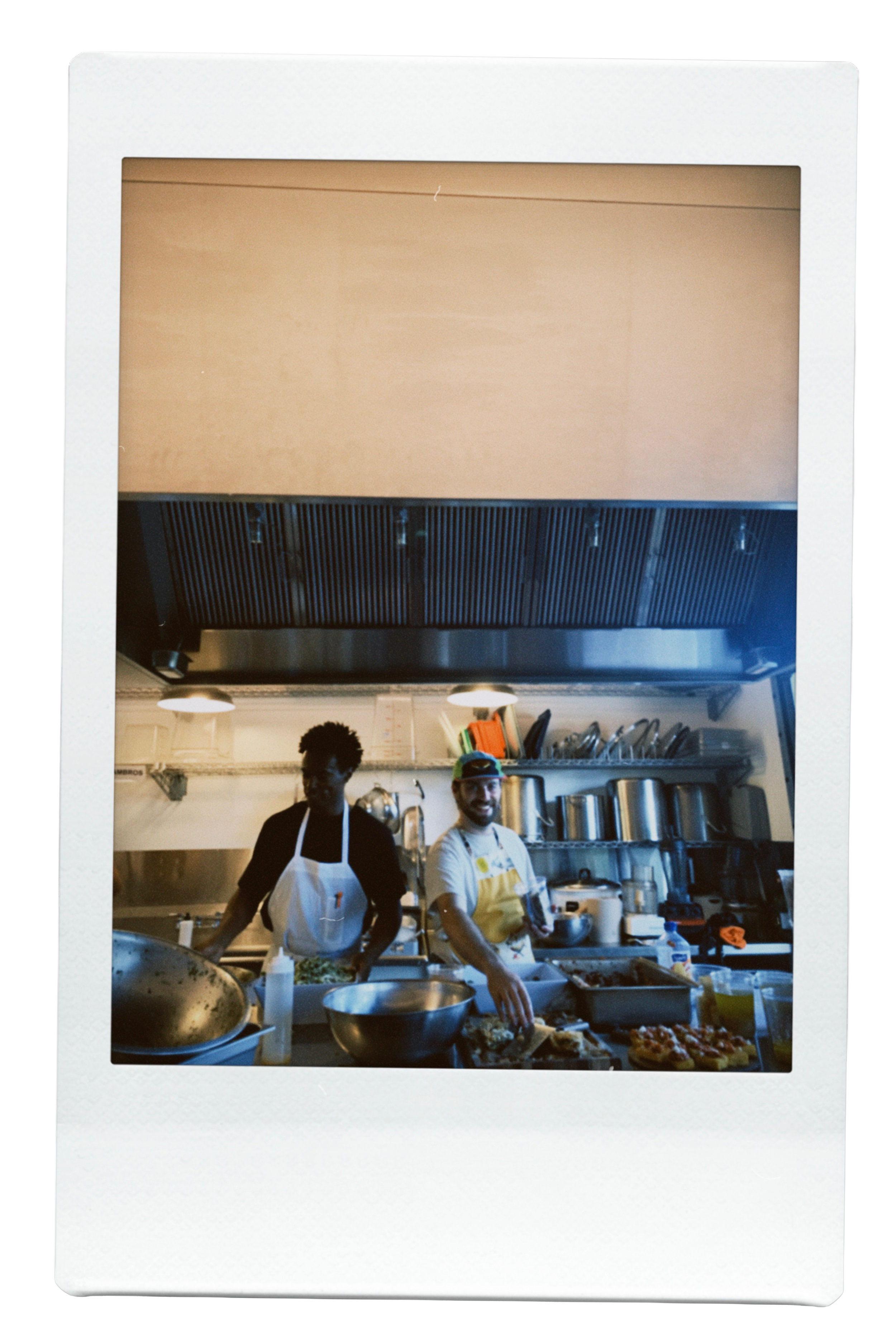 Two chefs working in a commercial kitchen, surrounded by cooking utensils and ingredients, smiling at the camera.