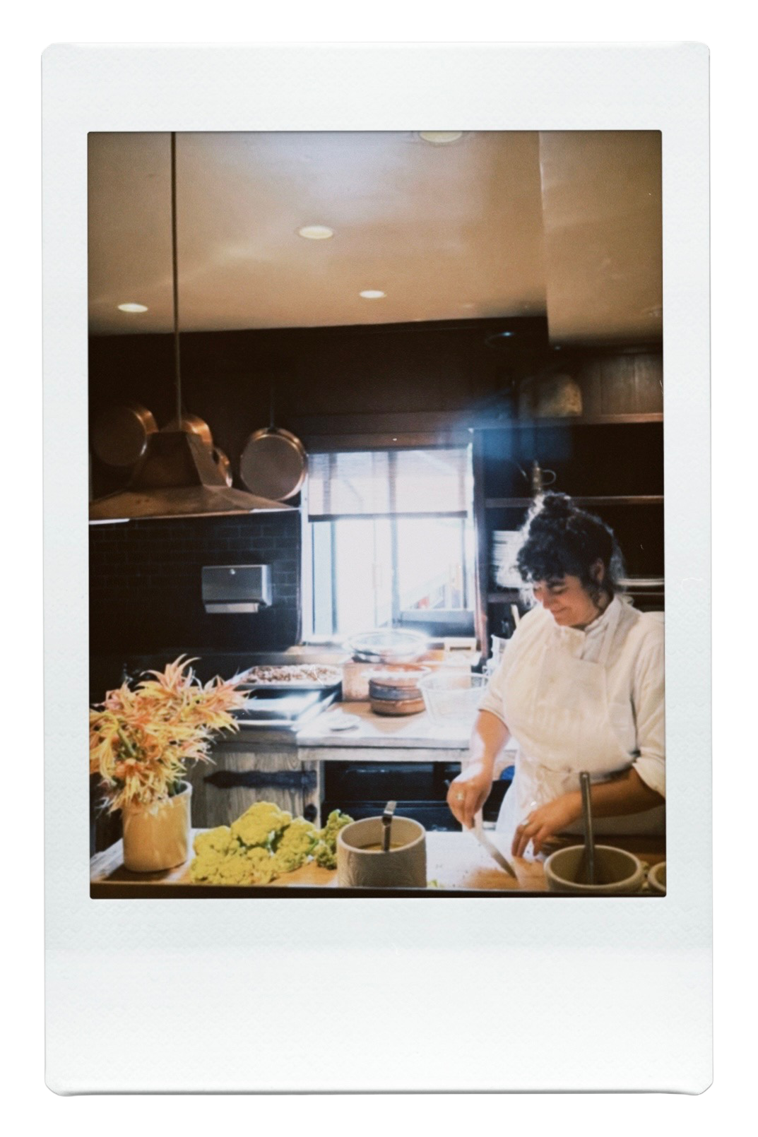 A woman preparing food in a kitchen with a large window, floral arrangement, and various kitchen utensils and supplies.