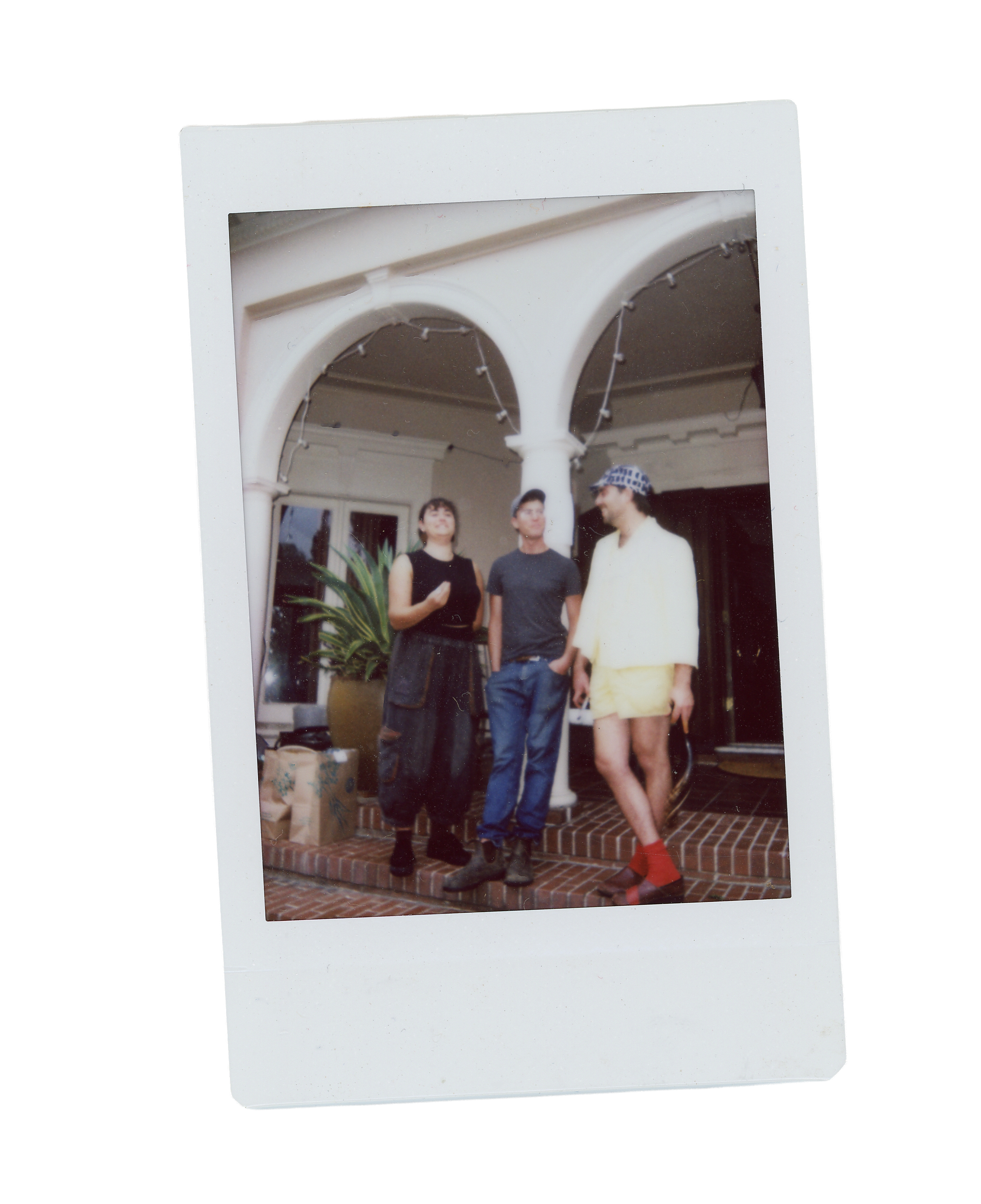 Three people standing on a brick porch under a white archway with string lights, including a woman with her hand up, a man in a dark T-shirt, and a person in a yellow outfit with red socks and a checkered hat.