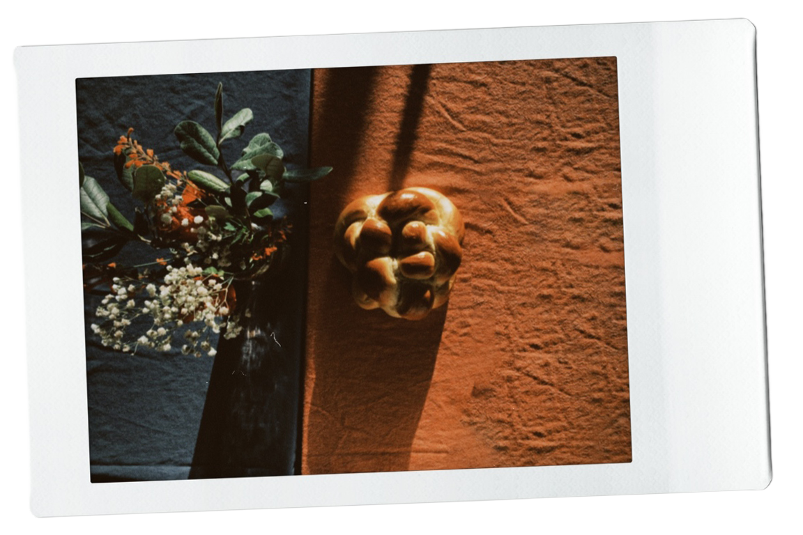An image of a small floral bouquet on dark cloth and a round challah on an orange-textured surface.
