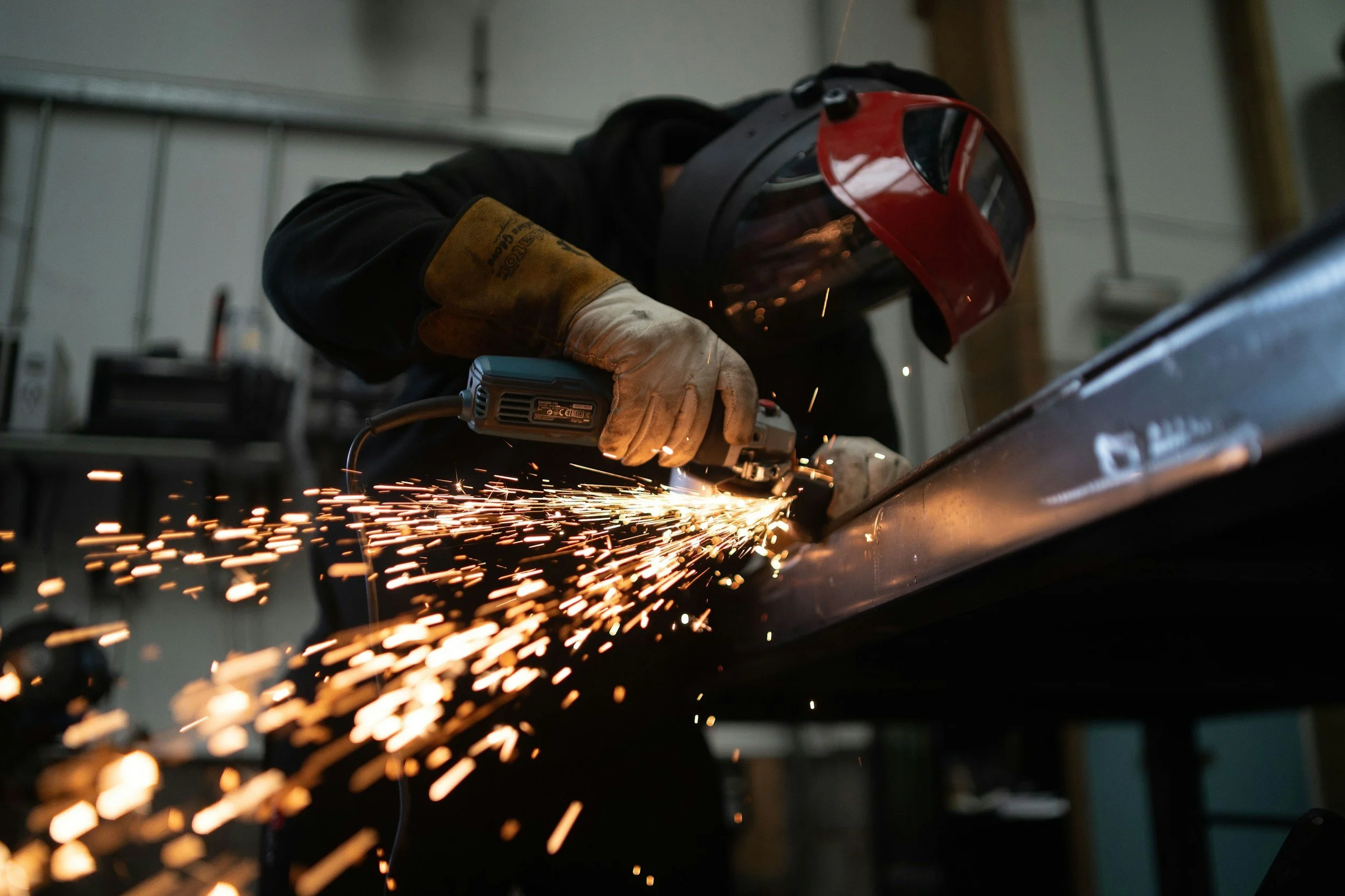 A person wearing protective gloves, a welding helmet, and dark clothing welding a metal piece in a workshop, with bright sparks flying from the welding process.