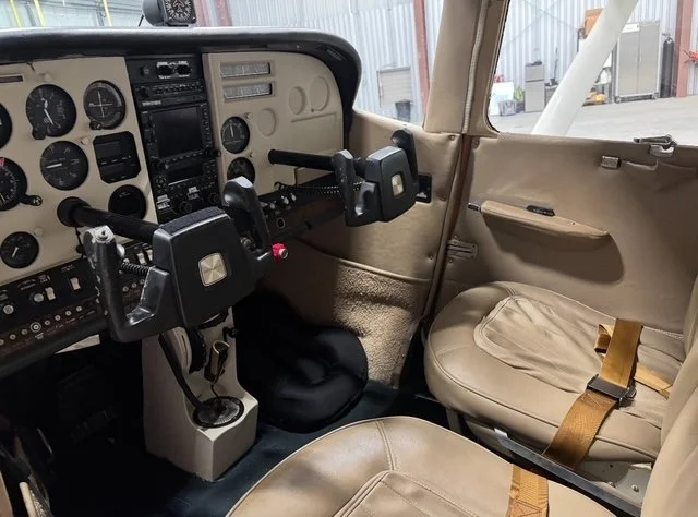 Interior cockpit of an aircraft showing control yokes, instrument panel, and beige pilot and passenger seats.