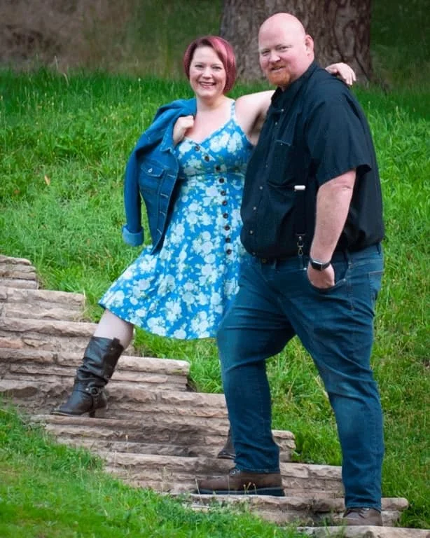 A woman with pink hair, wearing a blue and white flowered sundress stands with her arm across the shoulders of a bald man with a reg goatee. She is standing three steps higher up the stairs so they look closer in height