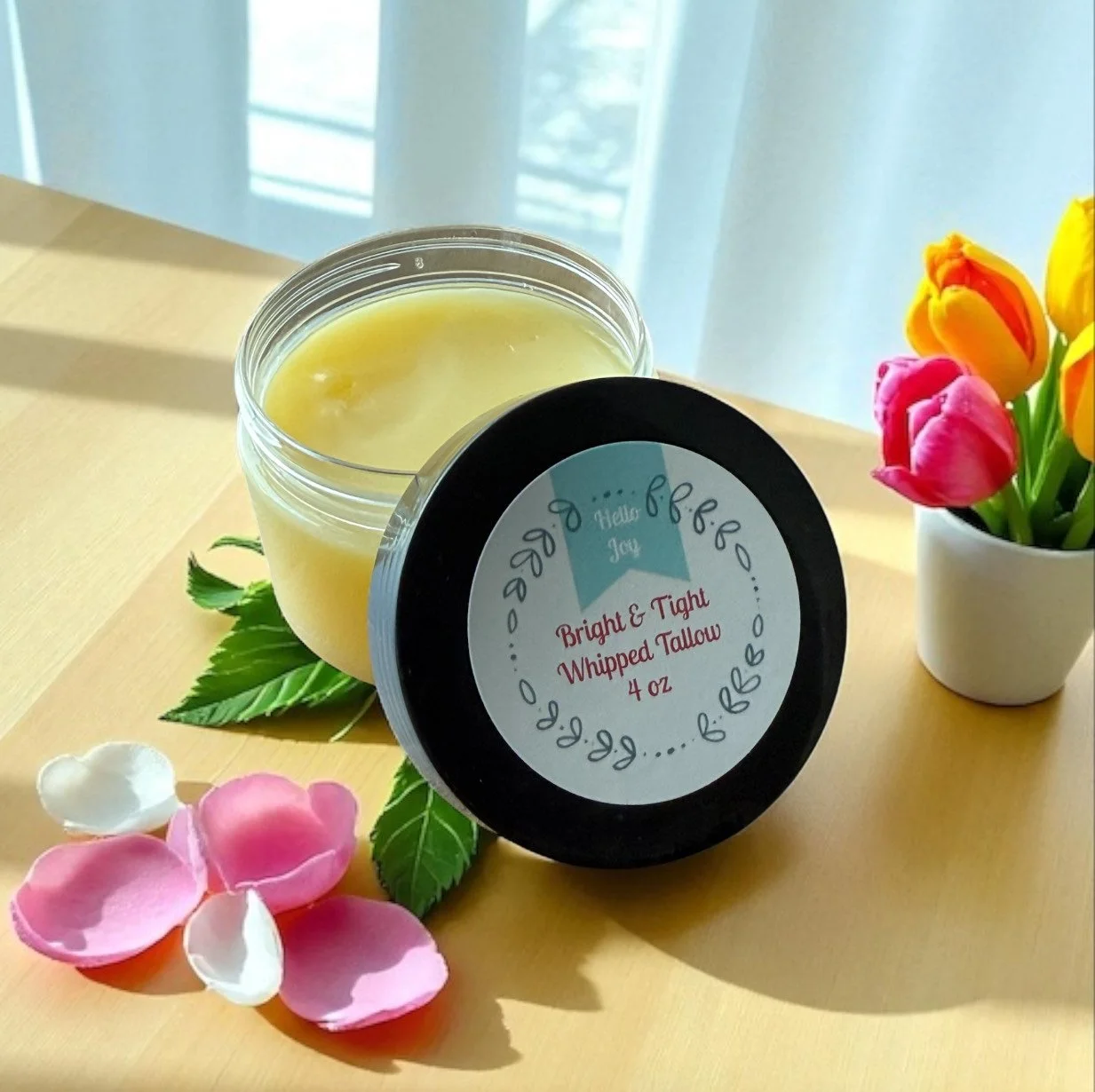 A jar of bright and tight whipped tallow labeled 4 oz with a lemon-colored candle inside, decorated with green leaves and pink flower petals, placed on a wooden surface next to a small vase of tulips with pink, yellow, and orange blooms.