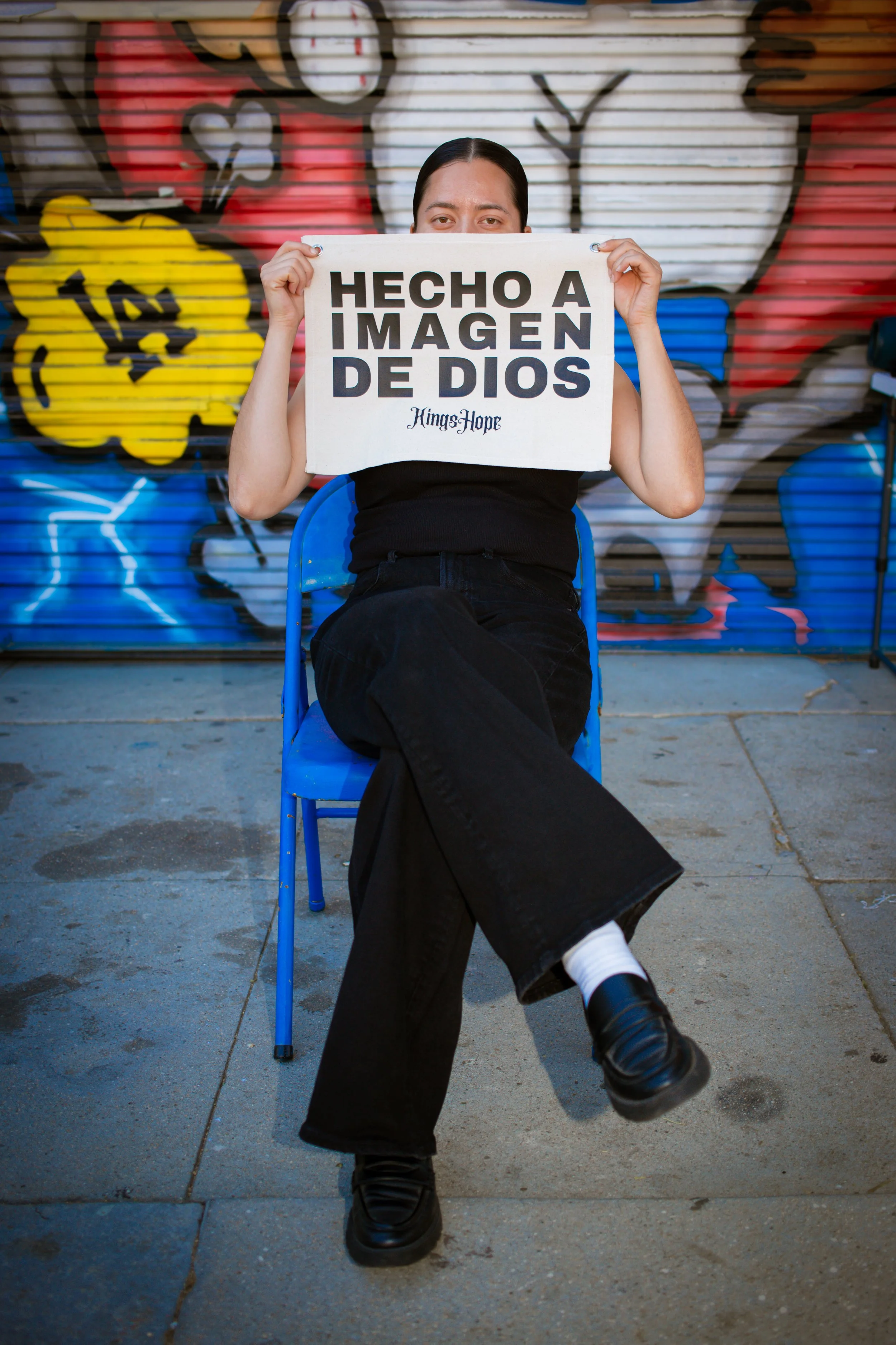 Woman holding KingsHope Hecho A Imagen De Dios canvas banner in front of East LA graffiti wall