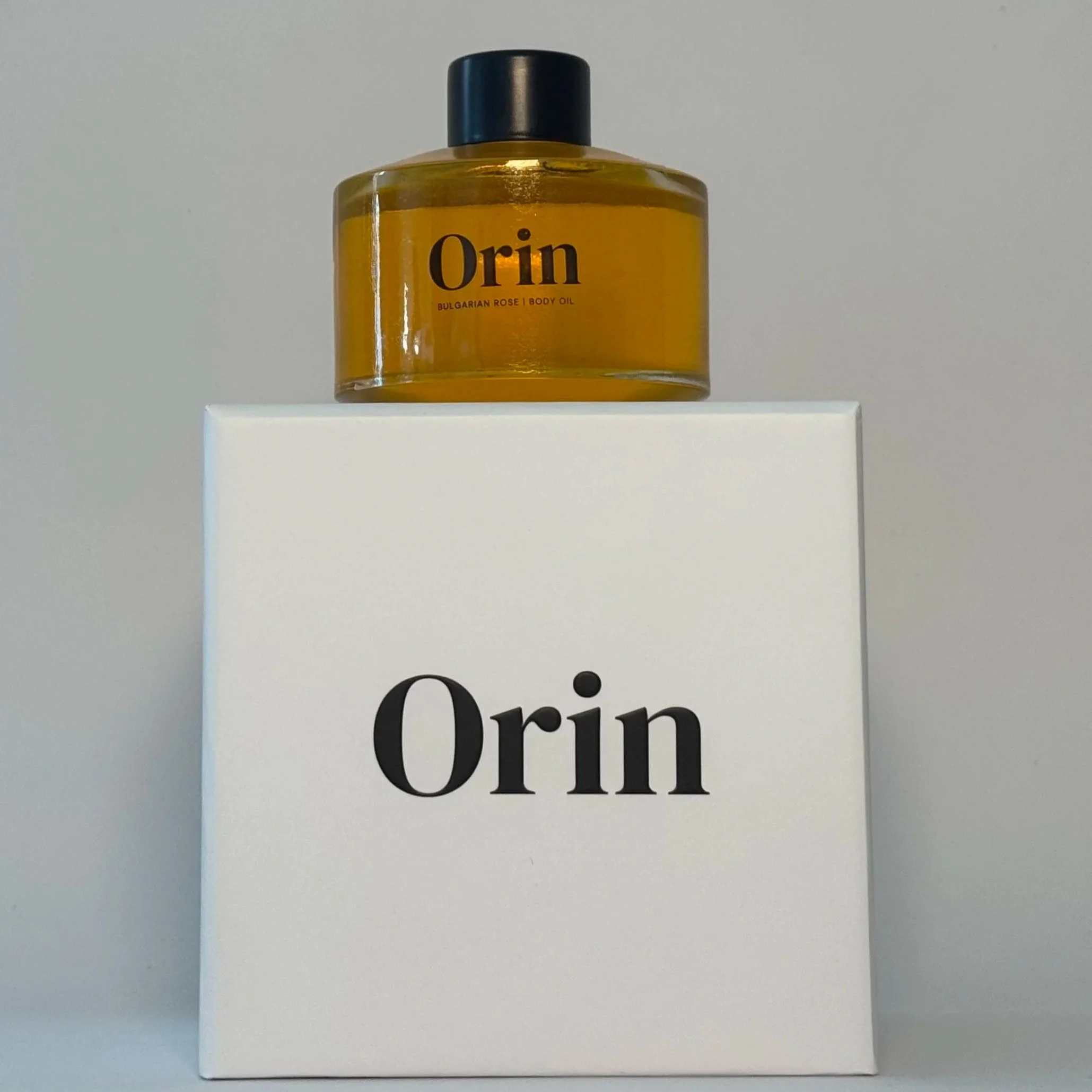 100ml glass bottle of Orin Bulgarian Rose Body Oil atop a white luxury box. The golden hue of this high-performance botanical skincare formula promotes skin longevity and lipid barrier support.