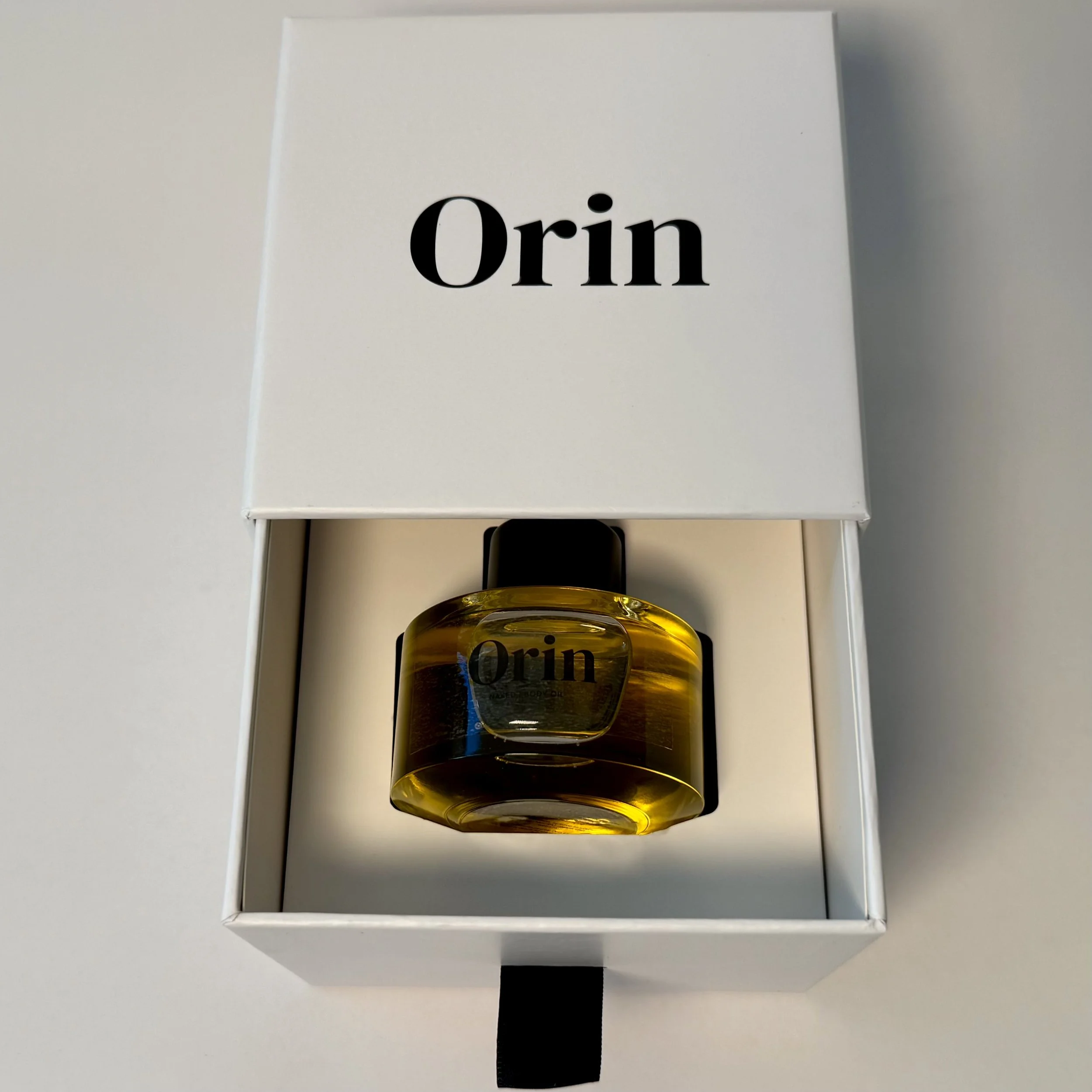 Top-down view of 50ml Orin Naked Body Oil in an open white luxury skincare box. The warm golden oil reflects the purity of this unscented ritual, perfect for targeted antioxidant protection.