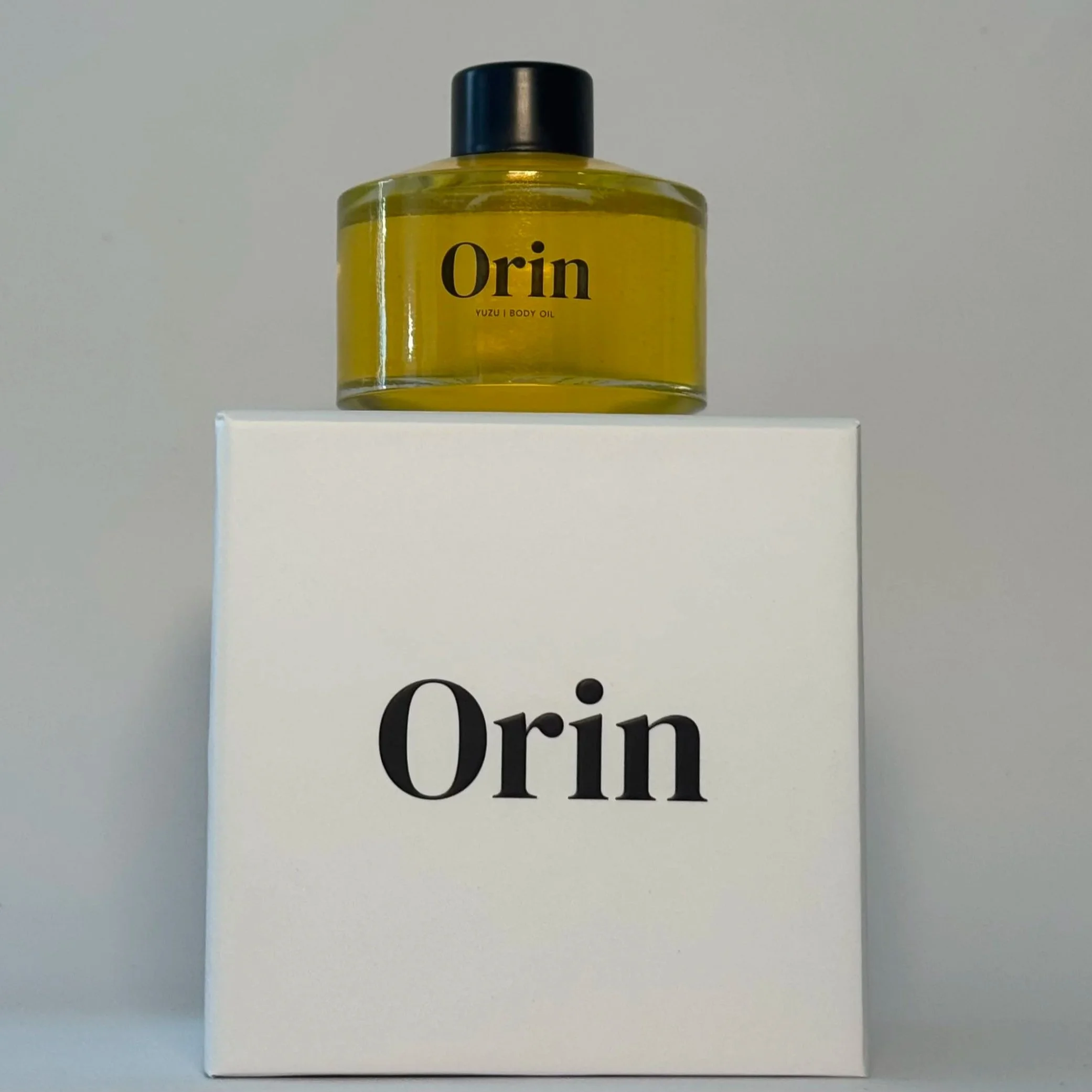 100ml glass bottle of Orin Yuzu Body Oil atop a white luxury box. The golden liquid showcases this energizing botanical formula, designed to promote skin luminosity and a surge of moisture.