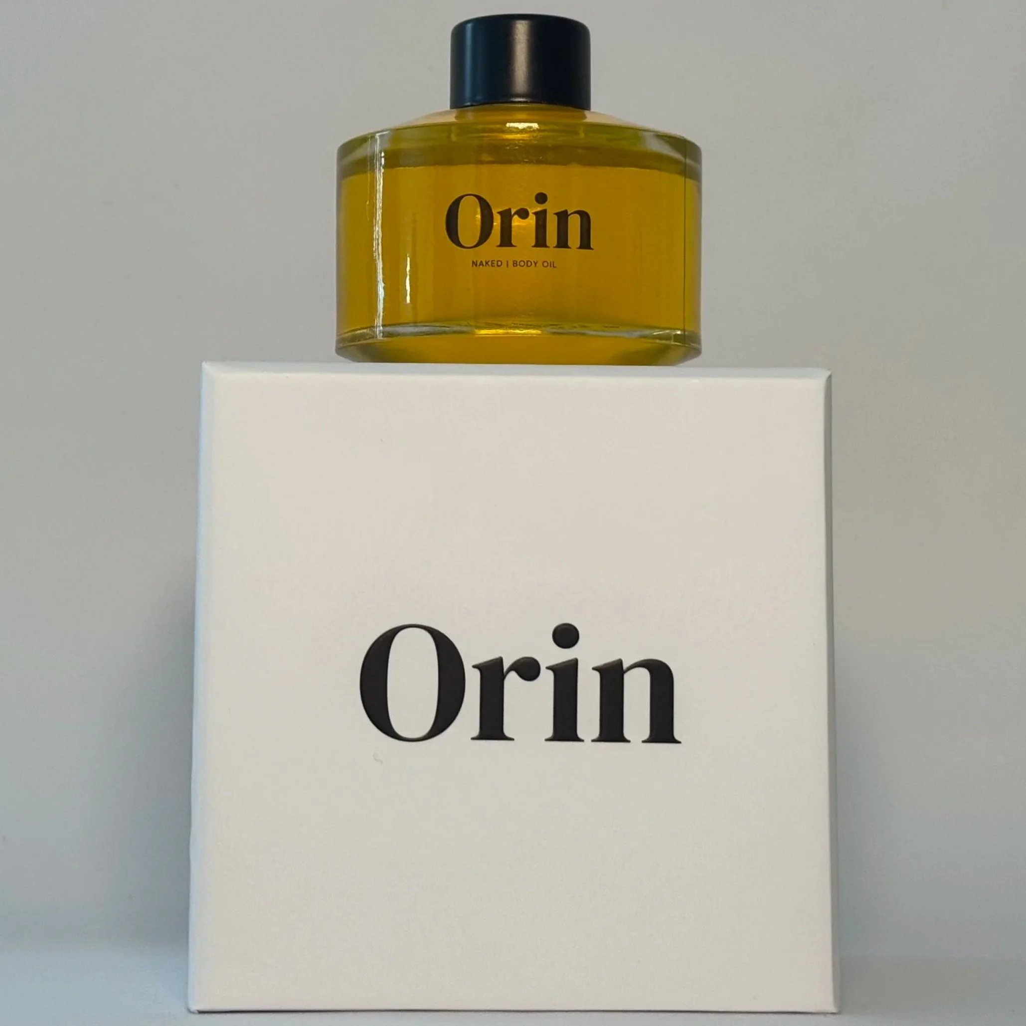 100ml glass bottle of Orin Naked Body Oil atop a white luxury box. The pure golden liquid highlights this unscented, fragrance-free botanical formula for lipid barrier support and resilience