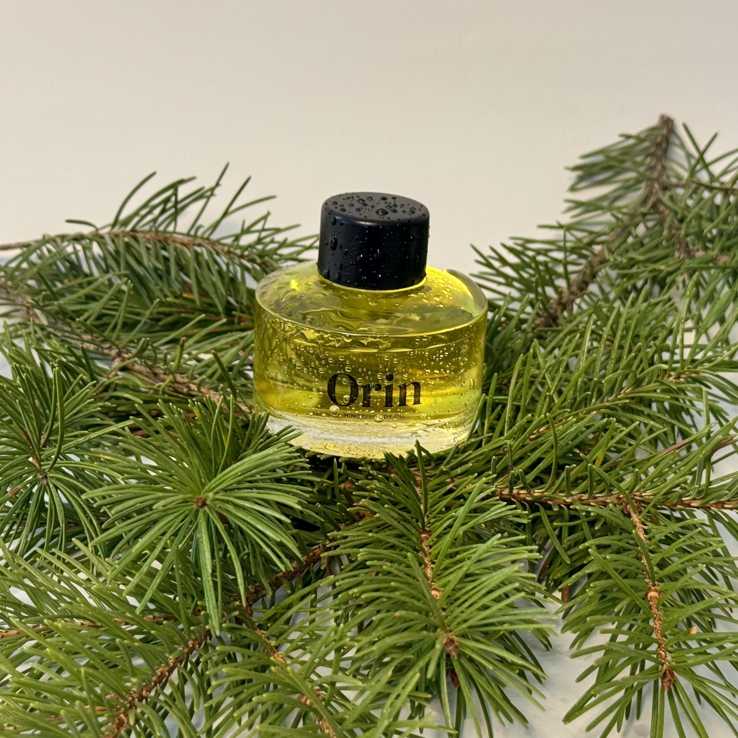Dewy Orin Hinoki Body Oil bottle on fresh pine branches. This natural setting highlights the Japanese Cypress and Omega-dense lipid blend designed for skin restoration and barrier support.