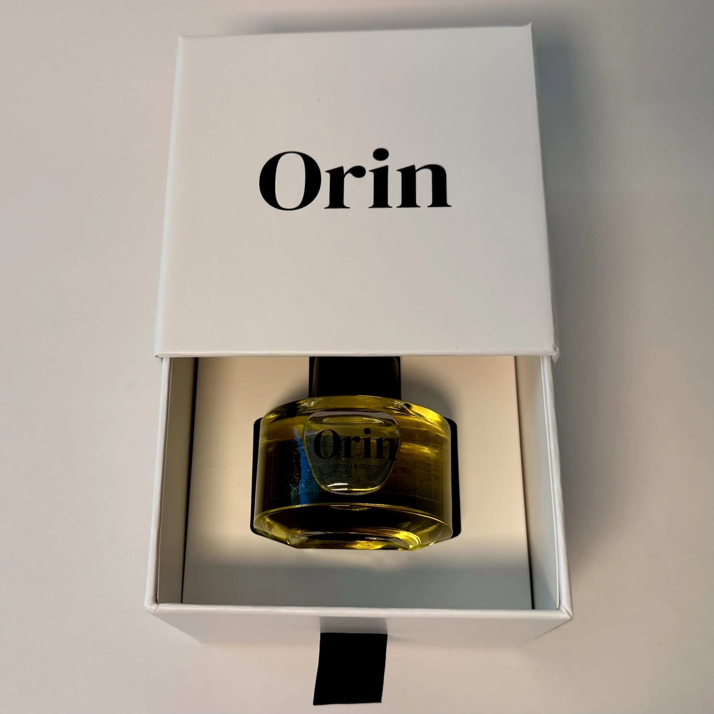 Top-down view of 50ml Orin Hinoki Body Oil in an open white luxury skincare box. The rich oil reflects the bio-nutrient foundation and restorative architecture of this lipid barrier treatment.