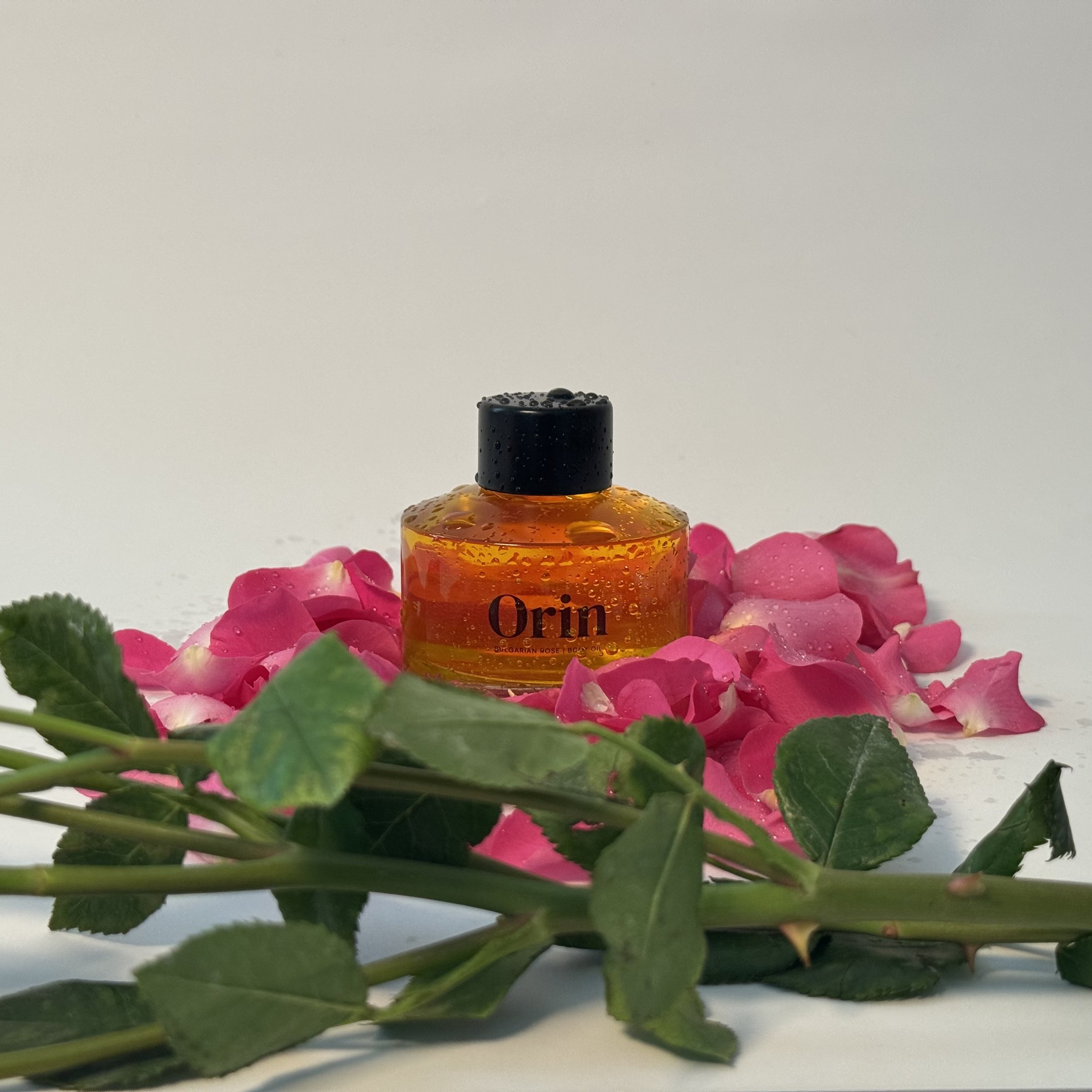 Orin Bulgarian Rose Body Oil bottle resting in pink rose petals and green stems. Dewy water droplets highlight this golden botanical oil, formulated for structural resilience and deep hydration.