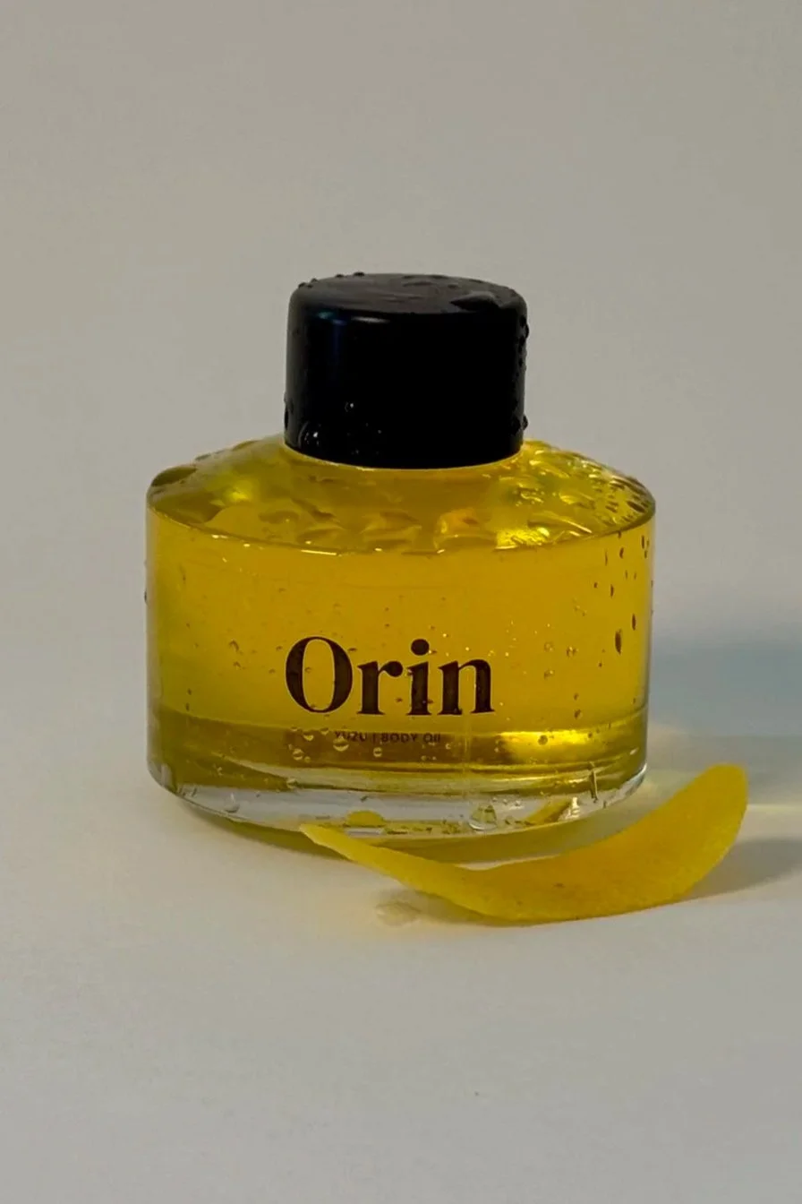 A 50ml glass bottle of Orin Yuzu Botanical Body Oil with a black cap, highlighting the brightening, energizing spring skincare benefits and ability to even skin tone.