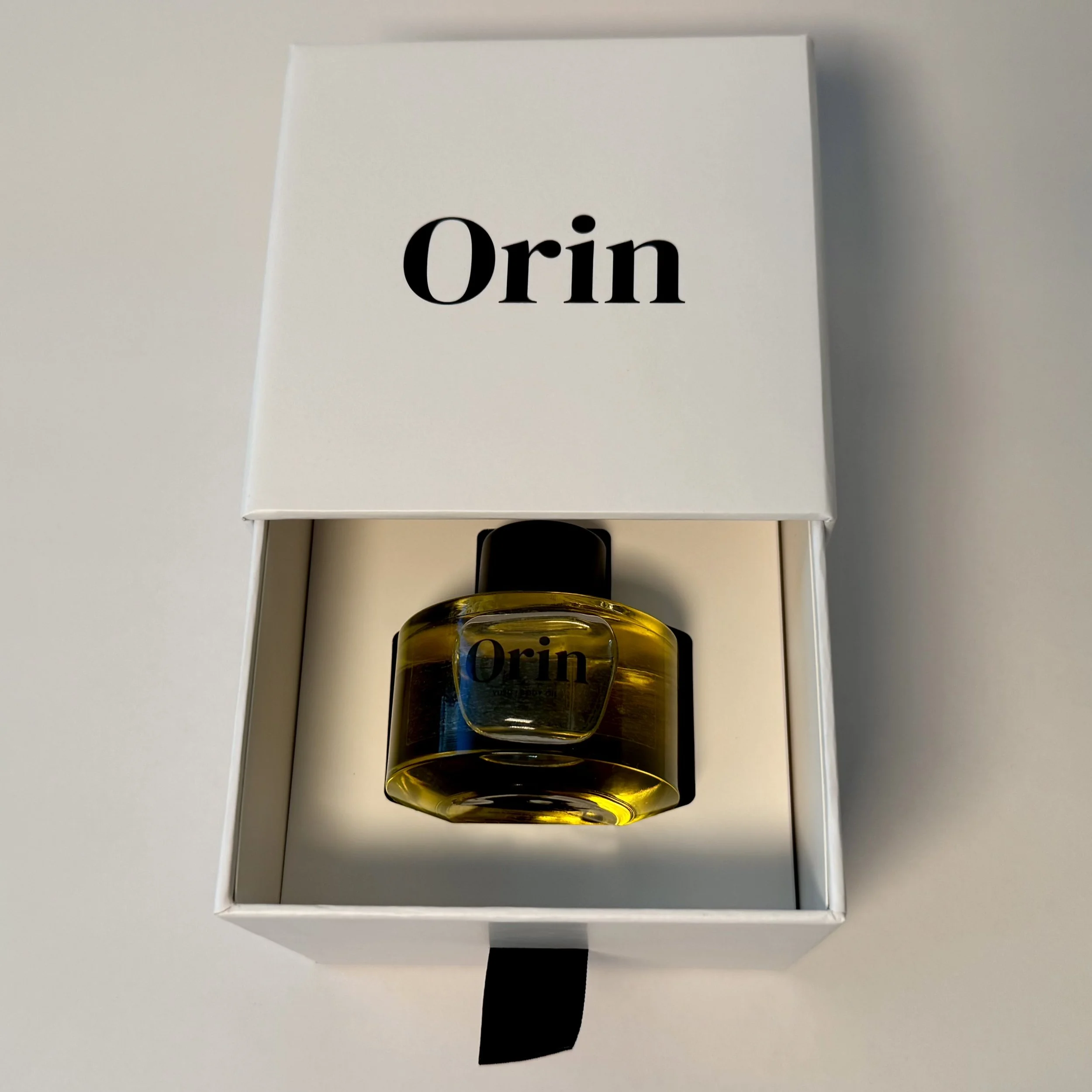 Top-down view of 50ml Orin Yuzu Body Oil in an open white luxury skincare box. The bright golden oil reflects the energizing botanical protection and awakening luminosity of this skincare ritual.