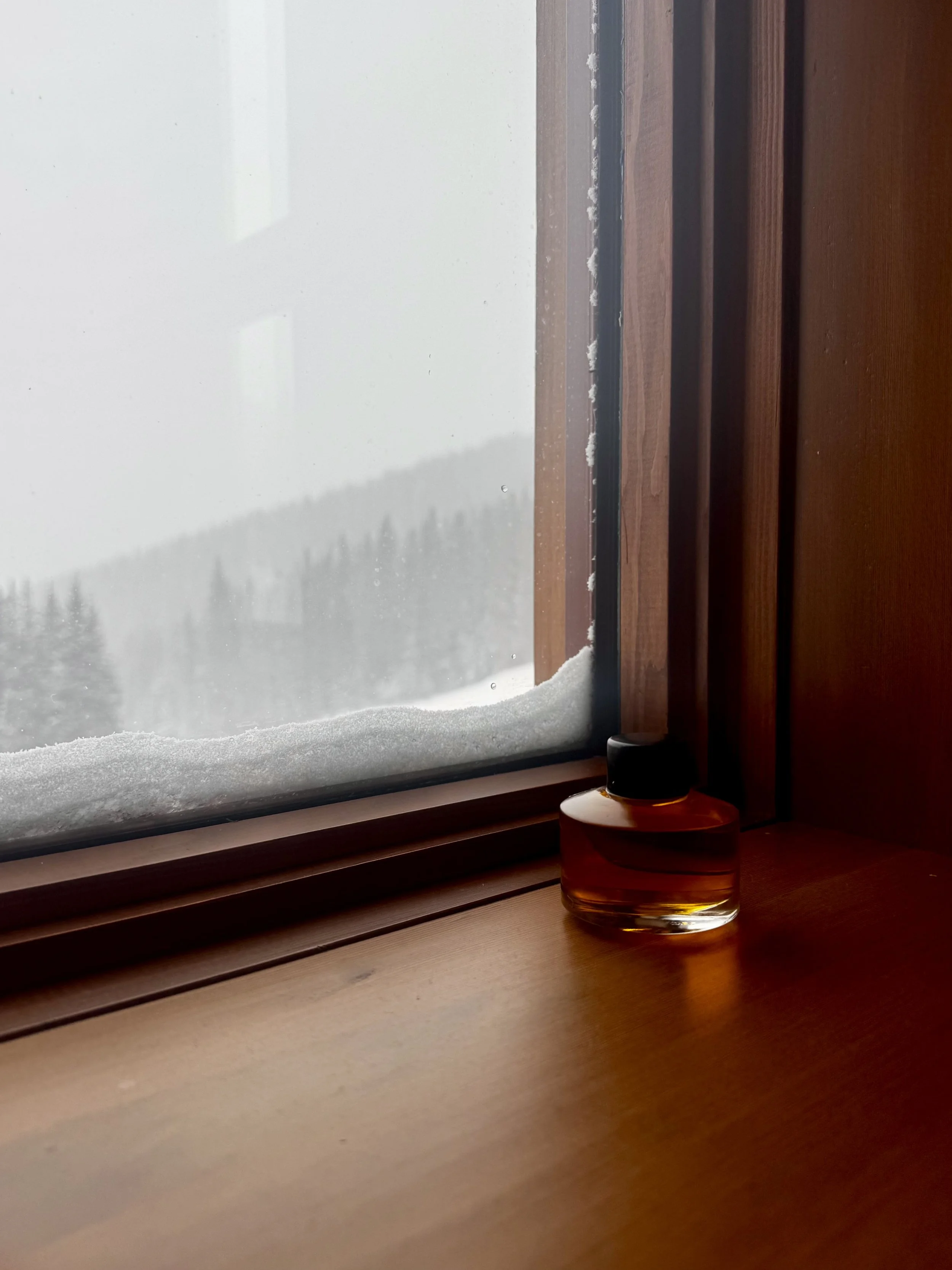 A glass bottle of amber-colored liquid on a wooden windowsill, with a snowy landscape and snow-covered trees visible outside the window.