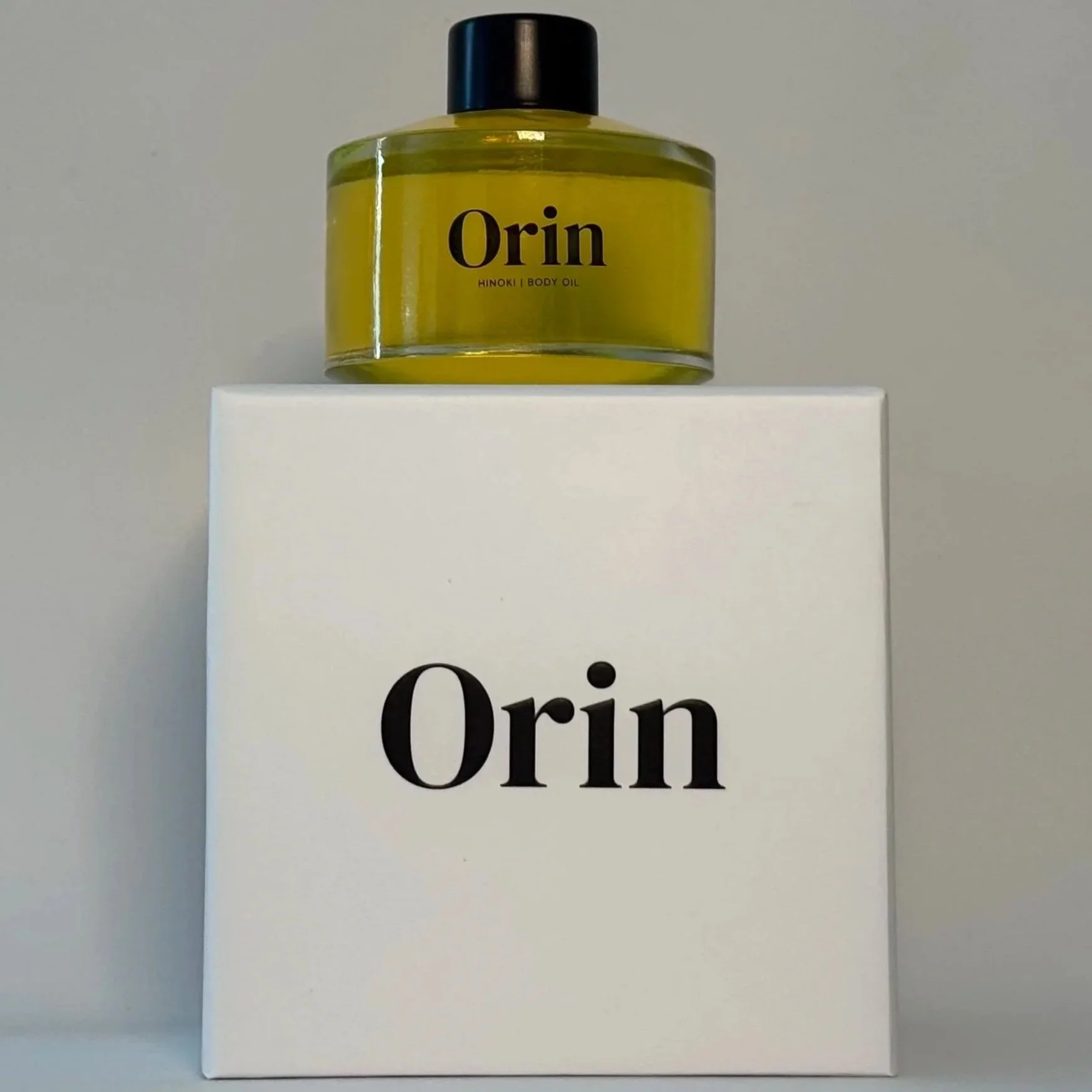 100ml glass bottle of Orin Hinoki Body Oil atop a white luxury box. The rich golden-green liquid showcases this high-performance botanical skincare formula for stability and restorative calm.