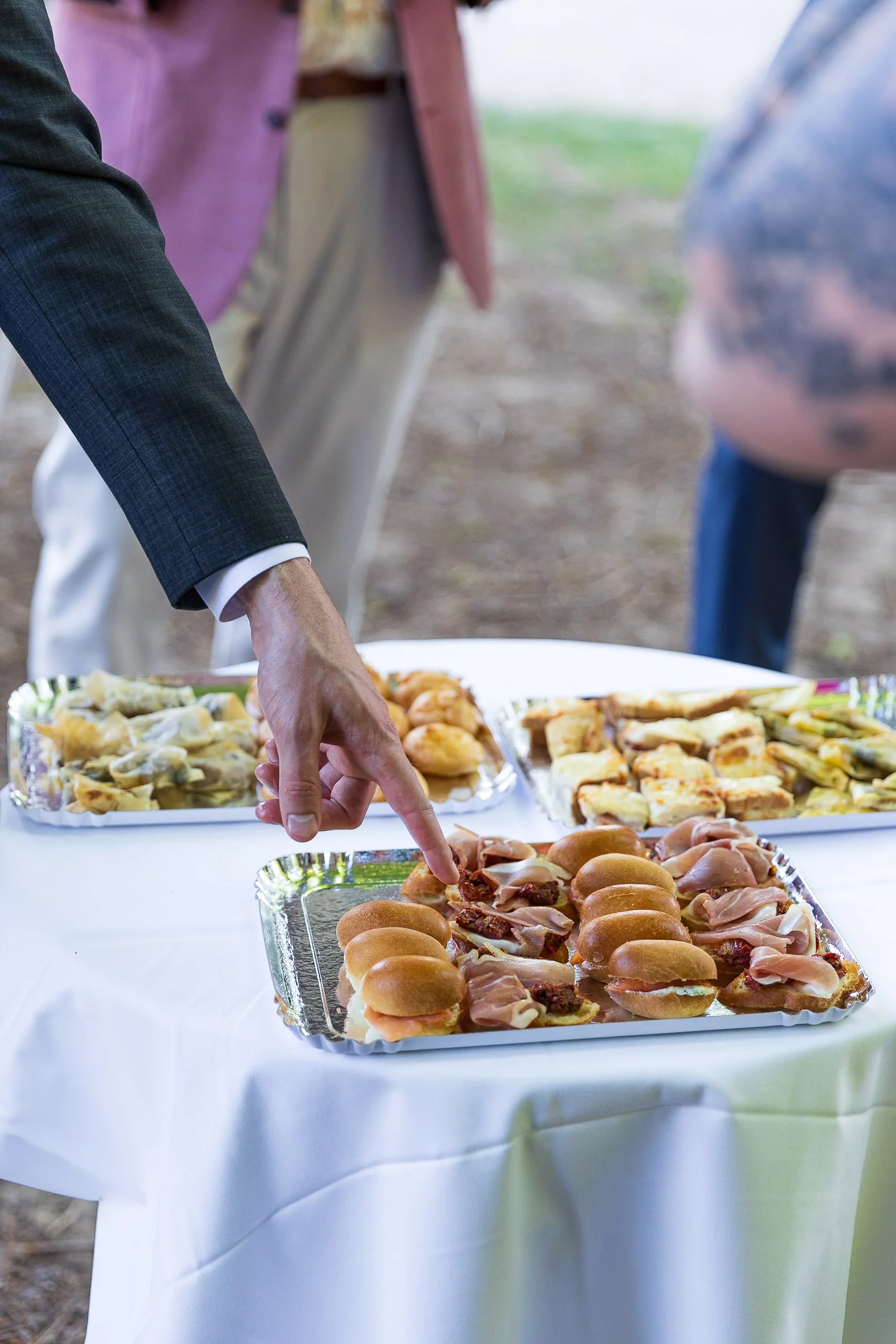 Close-up of a hand reaching for mini sliders on a tray at a buffet table with various finger foods.
