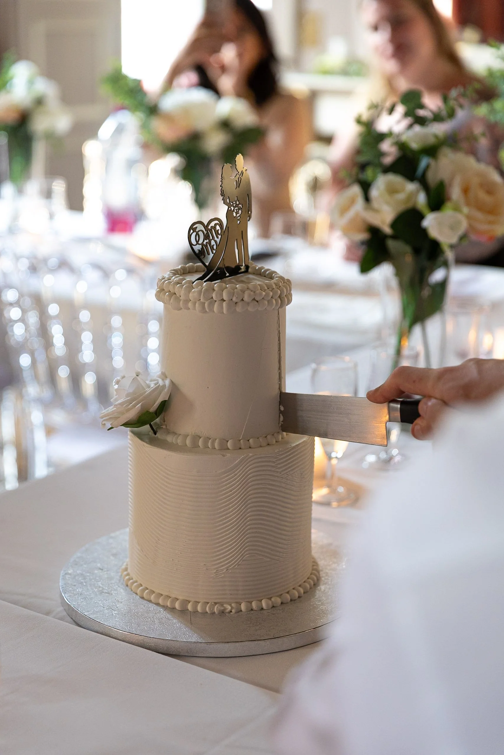 A wedding cake with a bride and groom topper, decorated with white icing and floral accents, situated on a silver base, set on a white tablecloth with a blurred background of people and floral arrangements at a wedding reception.