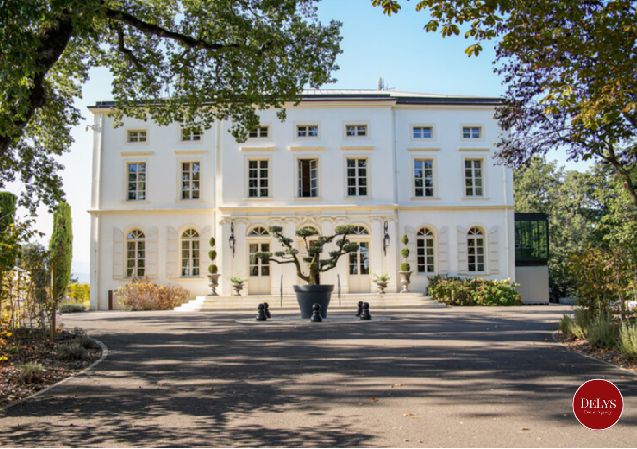 White multi-story mansion with multiple windows, large potted plants, and a central bonsai tree in a large pot outside on steps. Surrounded by trees and shrubbery. Logo reading "DeLys Event Agency" in the lower right corner.