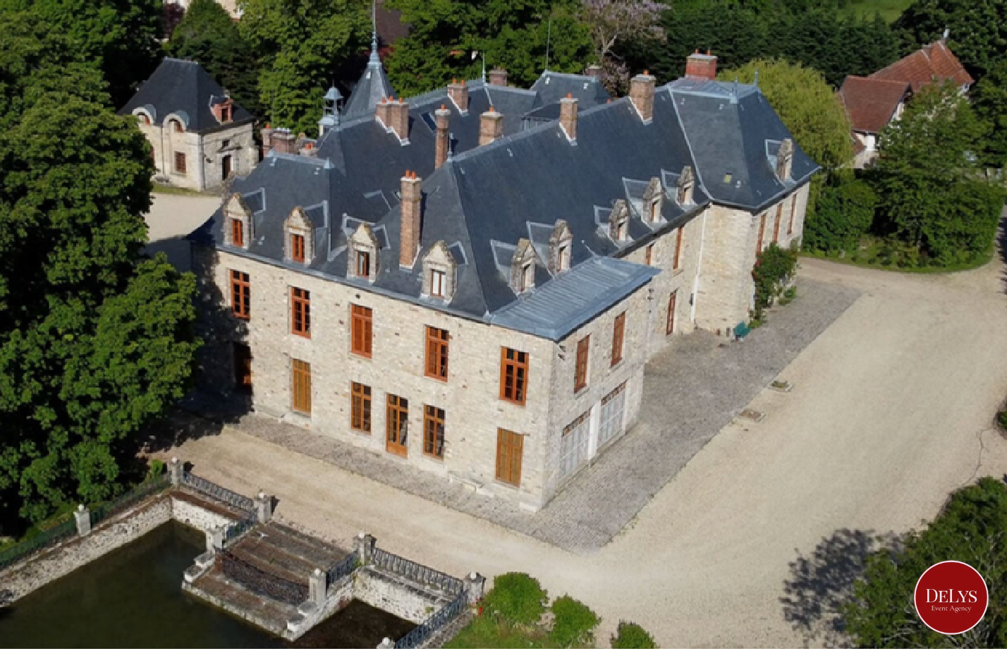 Aerial view of a historic stone mansion with a slate roof, surrounded by lush green trees and a gravel driveway.