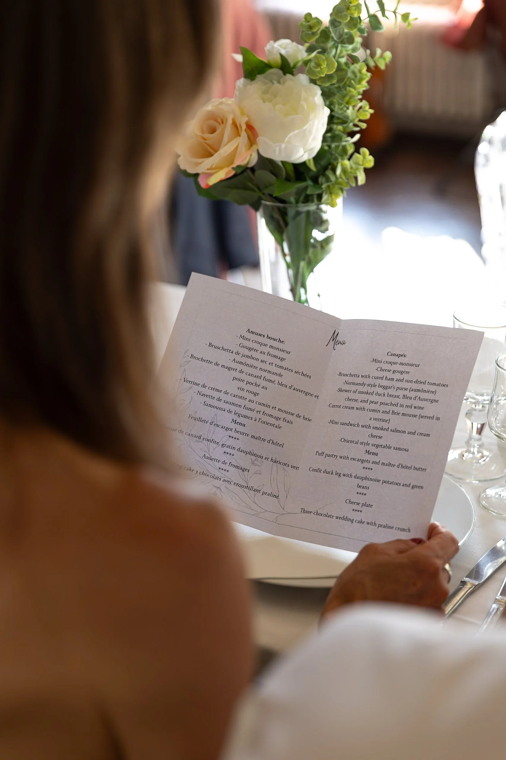 A person with brown hair holding a printed menu with French and English dishes, sitting at a table with a floral centerpiece featuring white roses and greenery, in a well-lit indoor setting.