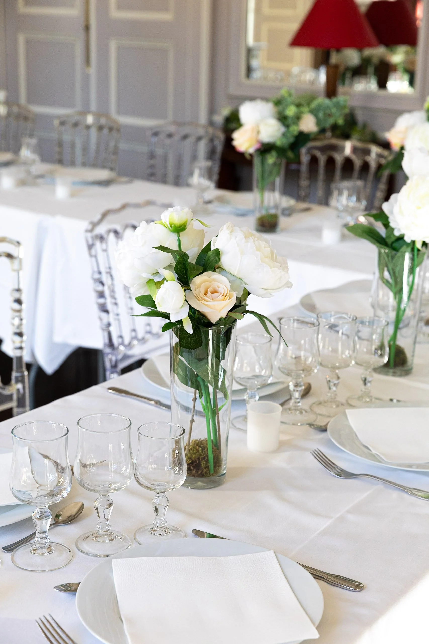 Elegant dining table set for a formal event with white tablecloths, glassware, white napkins, silverware, and floral centerpieces featuring white flowers and greenery in glass vases.