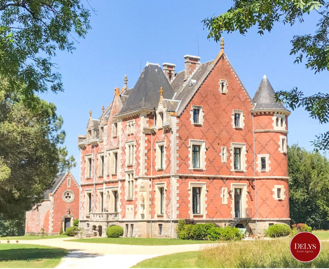 A large, historic castle with red brick and stone accents, multiple towers with conical roofs, surrounded by green trees and a grassy lawn, under a clear blue sky.
