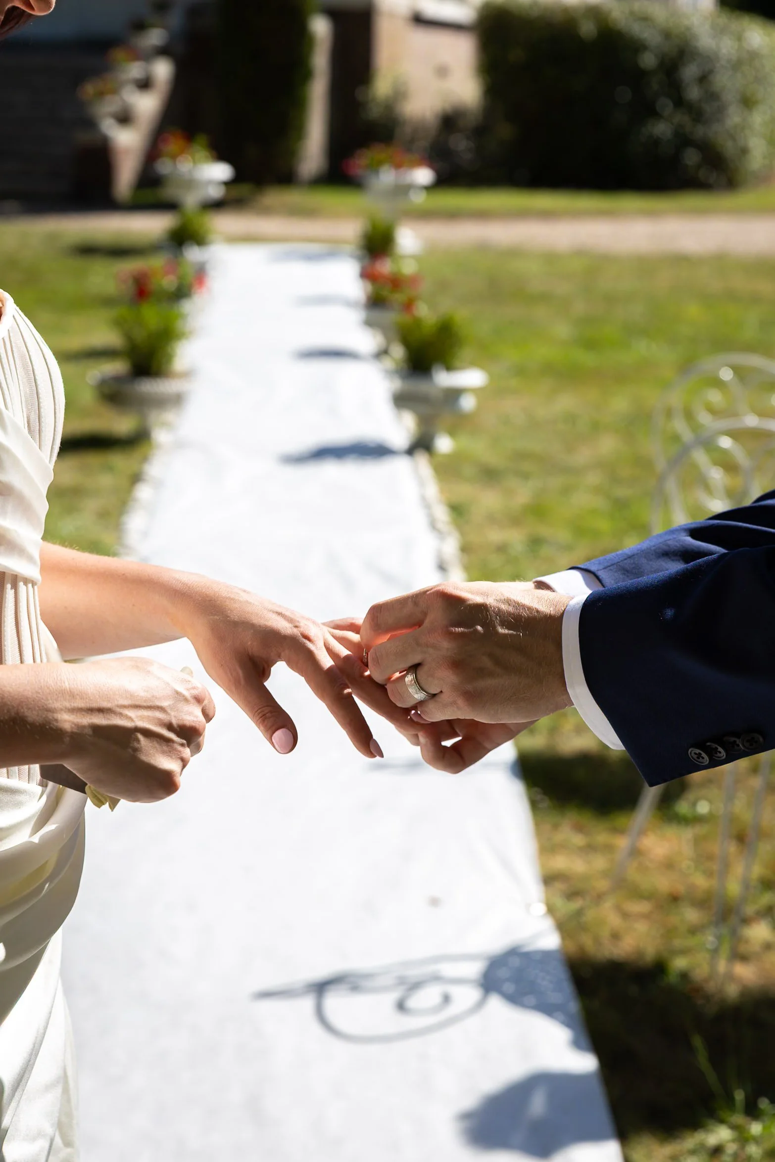 A close-up of a wedding ceremony where a bride and groom are holding hands, with the groom's wedding ring visible, outdoors with decorative flower arrangements in the background.