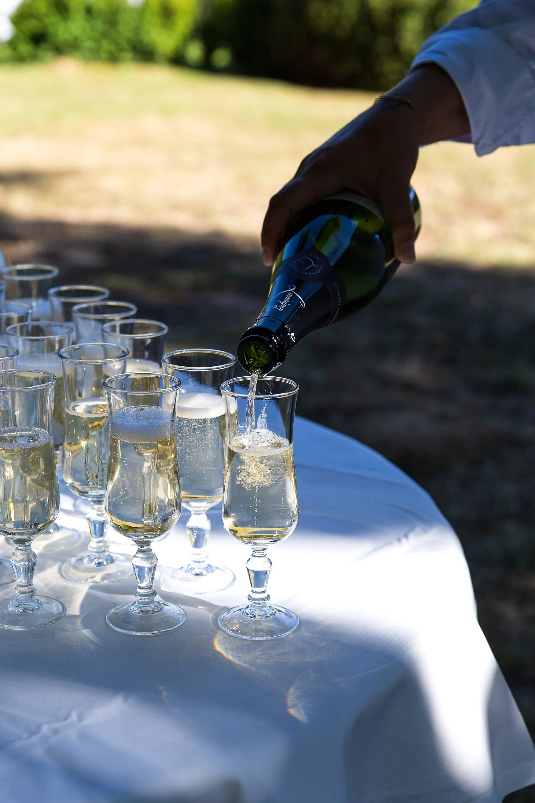 Person pouring champagne into a glass at an outdoor celebration with several filled champagne flutes on a white tablecloth.