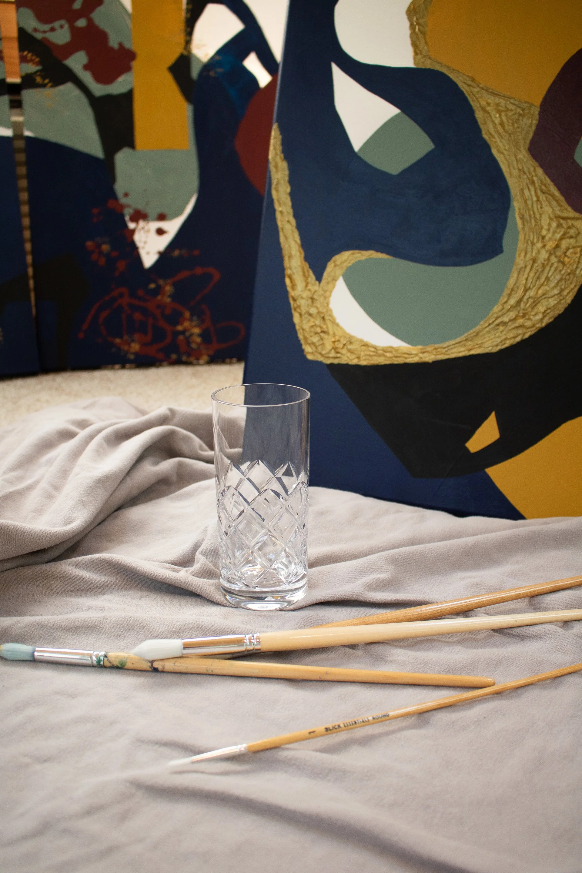 Painting brushes and a glass of water arranged in front of an original abstract painting from the Moments in Grief collection by Minnesota artist Nailah Ali.