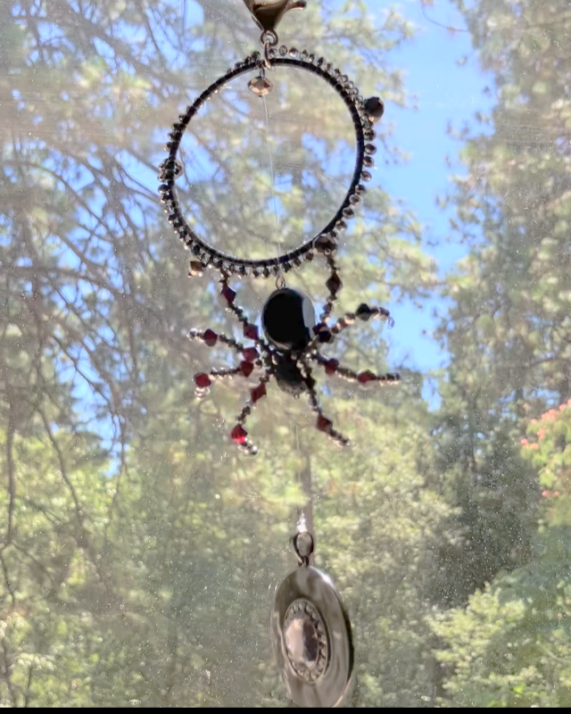 A crystal beaded spider hanging in a sunny window, with trees in the background, catching light.