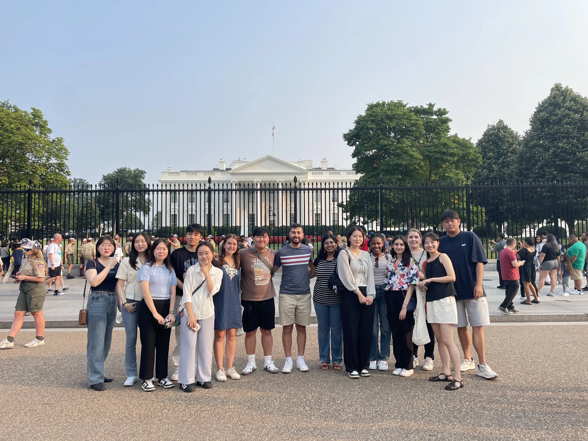 Group of diverse young adults posing in front of the White House with a fence and trees, some people in the background walking around and taking photos.