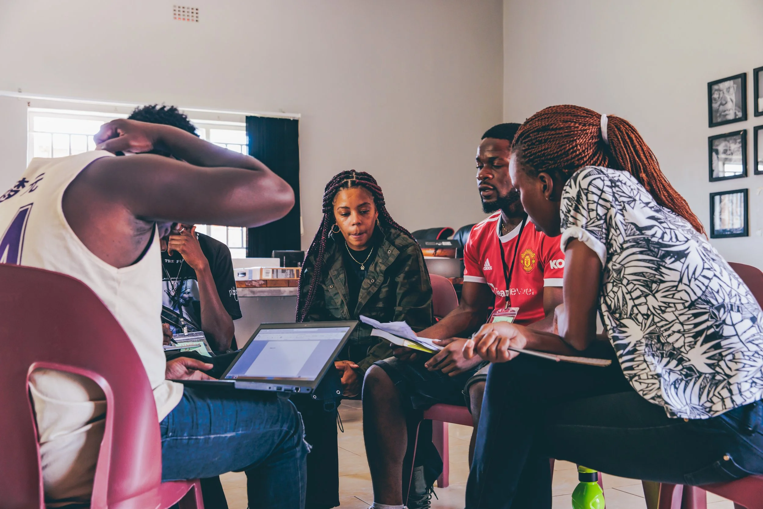 Group of five people sitting and discussing, with laptops and notebooks, in a room with photos on the wall.
