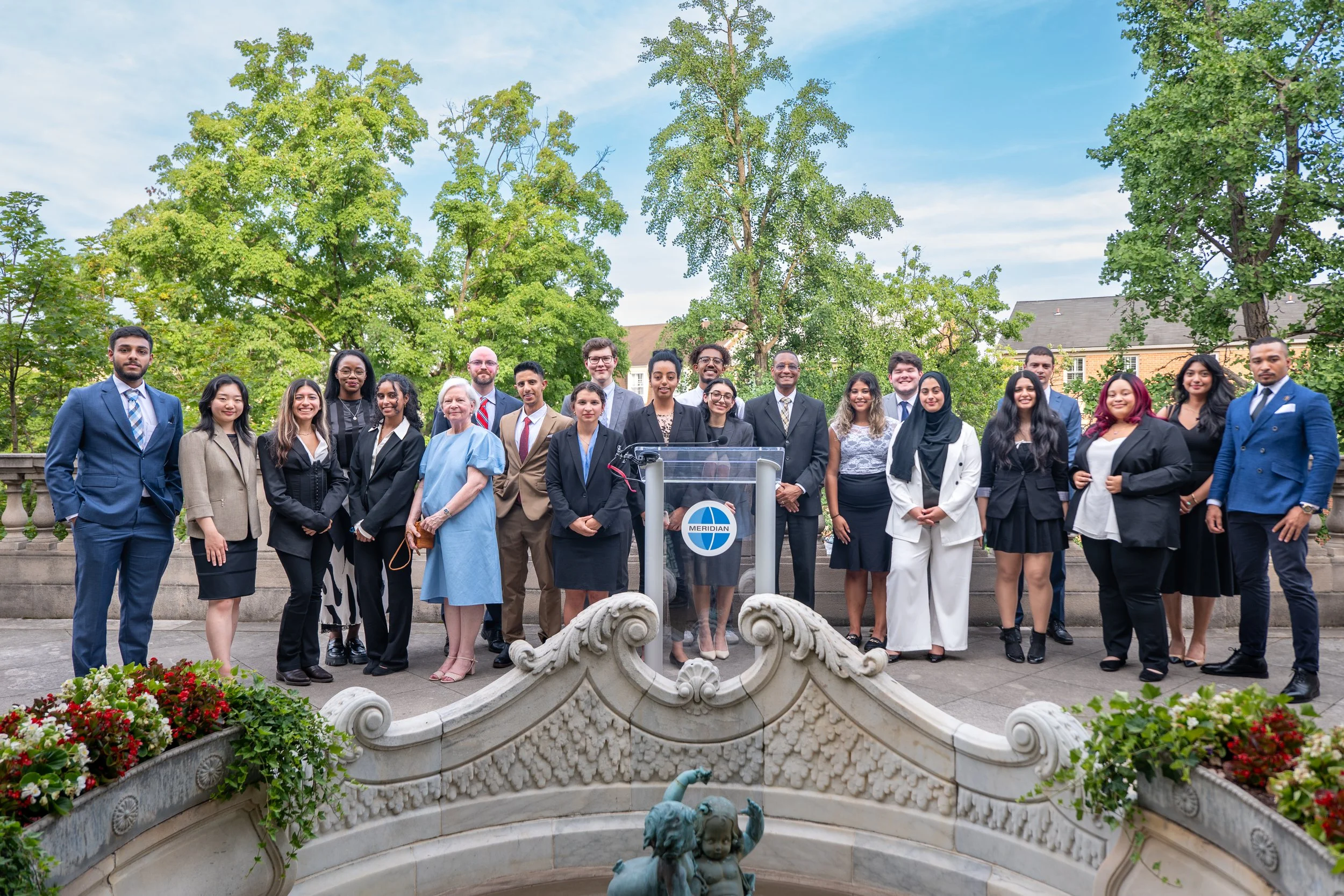 A diverse group of people posing outdoors in front of trees, composed of men and women in business attire, standing on a balcony. There is a fountain with a sculpture in the foreground and a sign with the word 'Meridian' on a stand. Clear skies are v